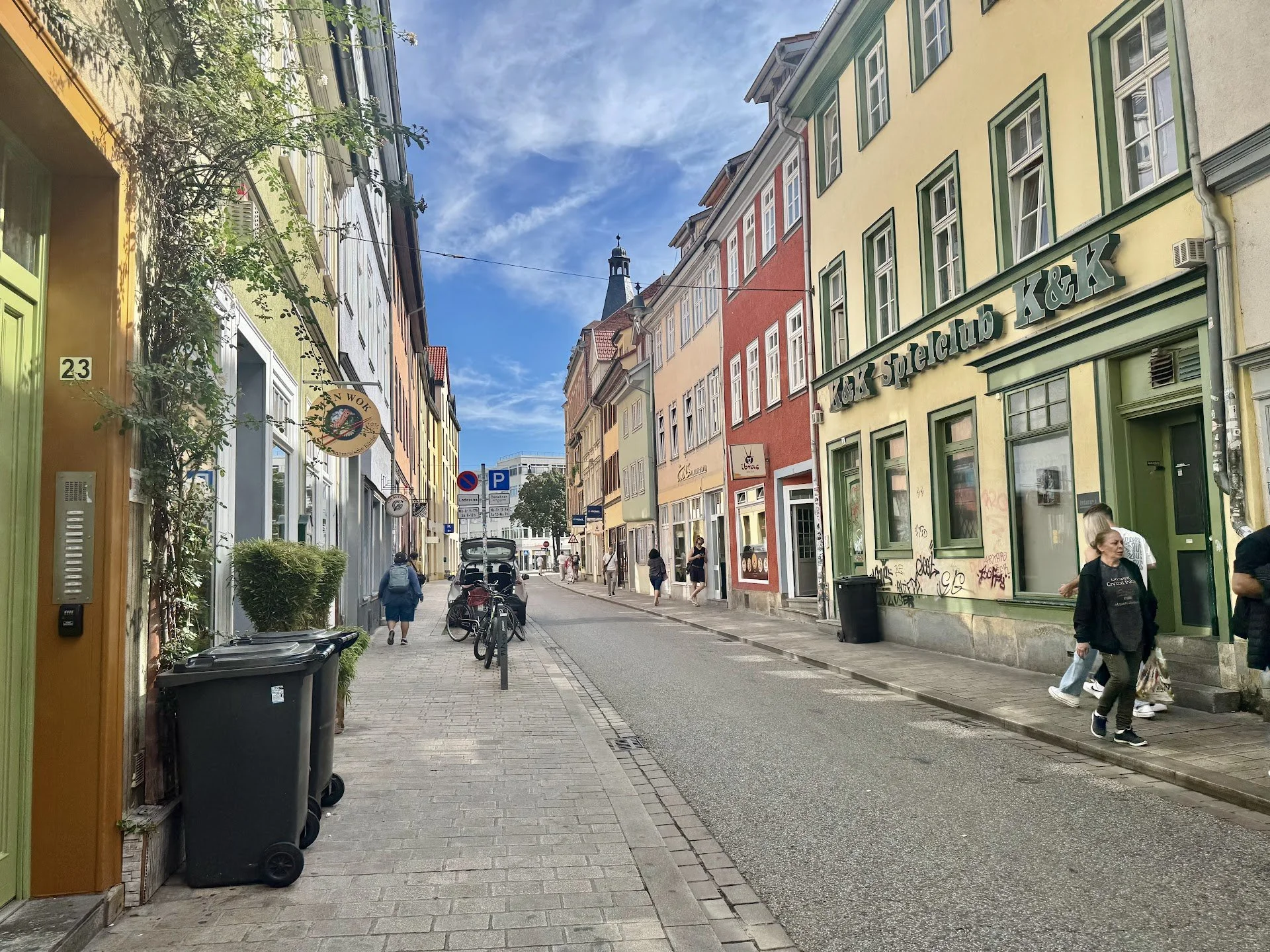 A narrow city street lined with colorful buildings, some with graffiti, and a church steeple in the background under a blue sky with wispy clouds. Pedestrians walk along the sidewalk, and bicycles are parked along the street.