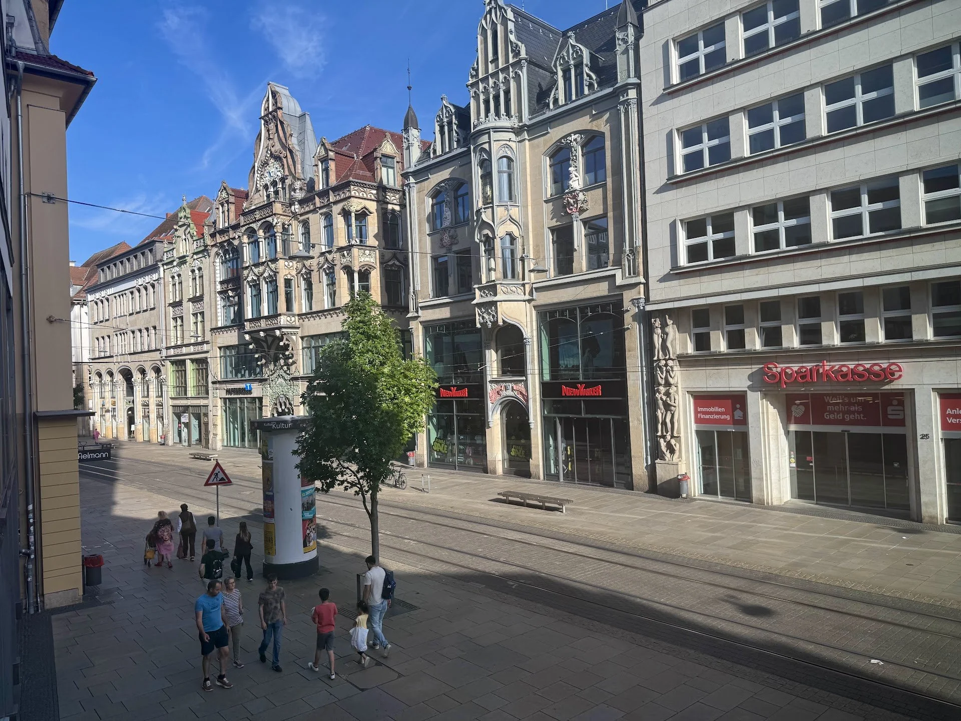 A city street with a mix of historic and modern buildings, pedestrians walking on the sidewalk, tram tracks, and a clear blue sky.