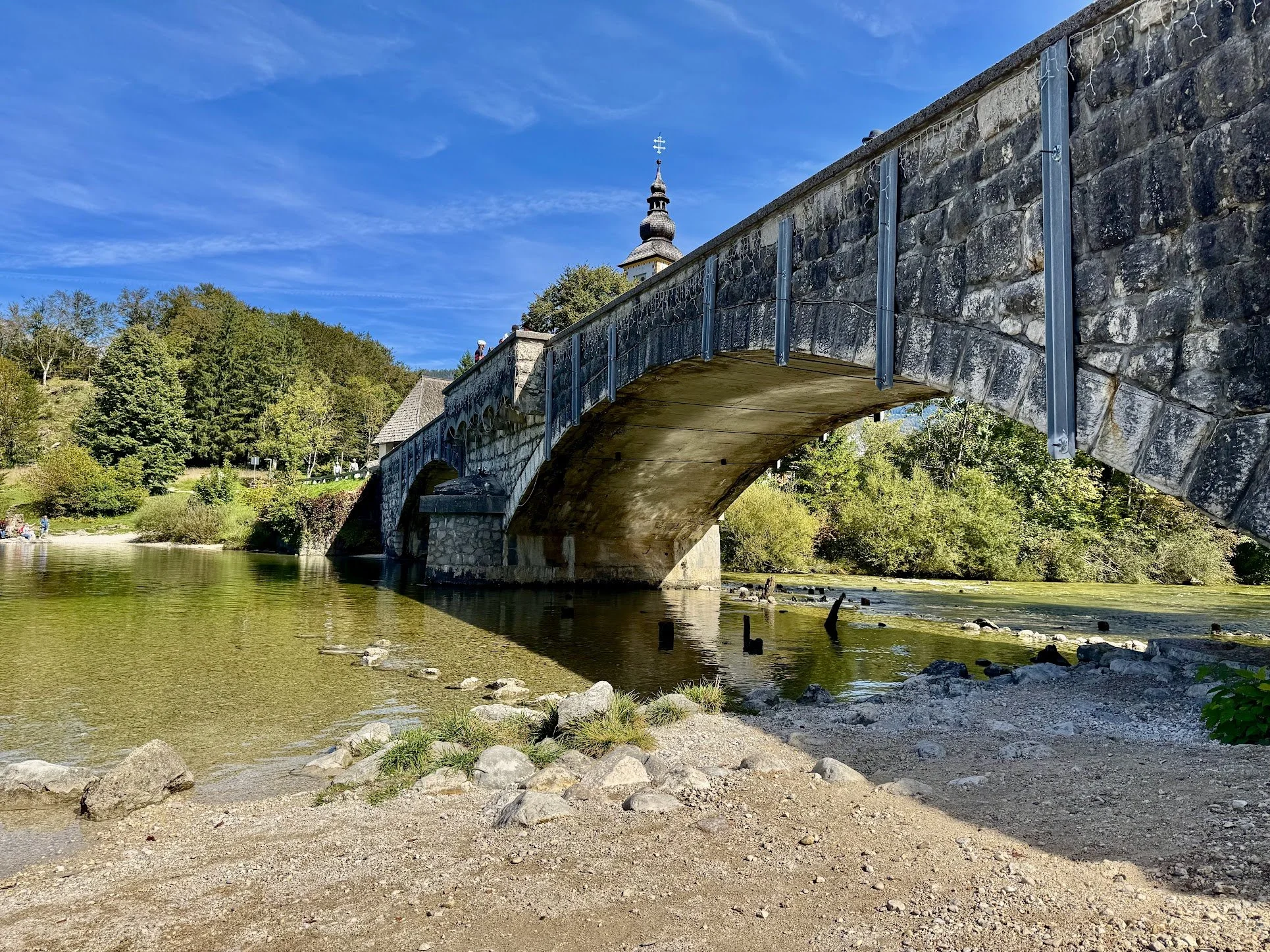 A stone pedestrian bridge over a river with a church steeple in the background, surrounded by green trees and a bright blue sky.