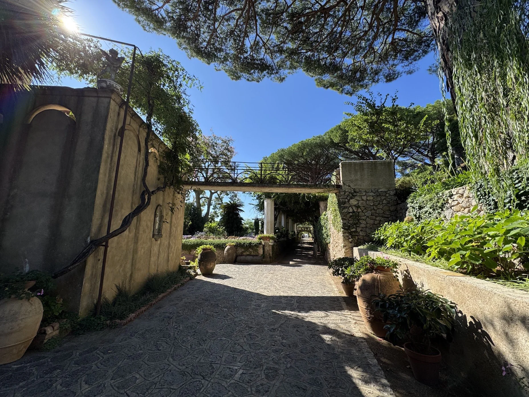 A charming outdoor garden pathway shaded by trees with a stone-paved walkway, large potted plants, and historic stone and stucco walls, under a clear blue sky with sunlight filtering through the branches.