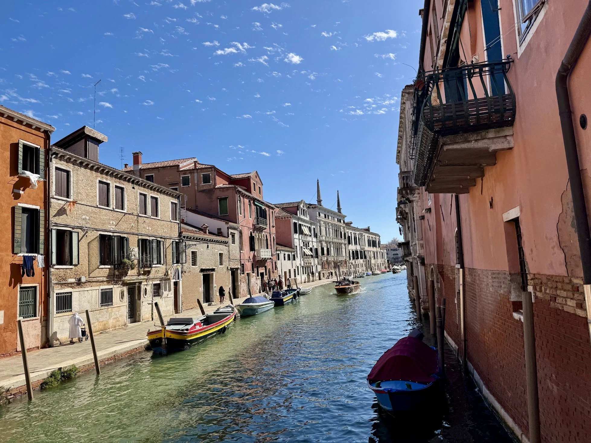 View of a canal with several small boats docked along the sides, colorful residential buildings with windows and balconies line the canal, under a partly cloudy blue sky.