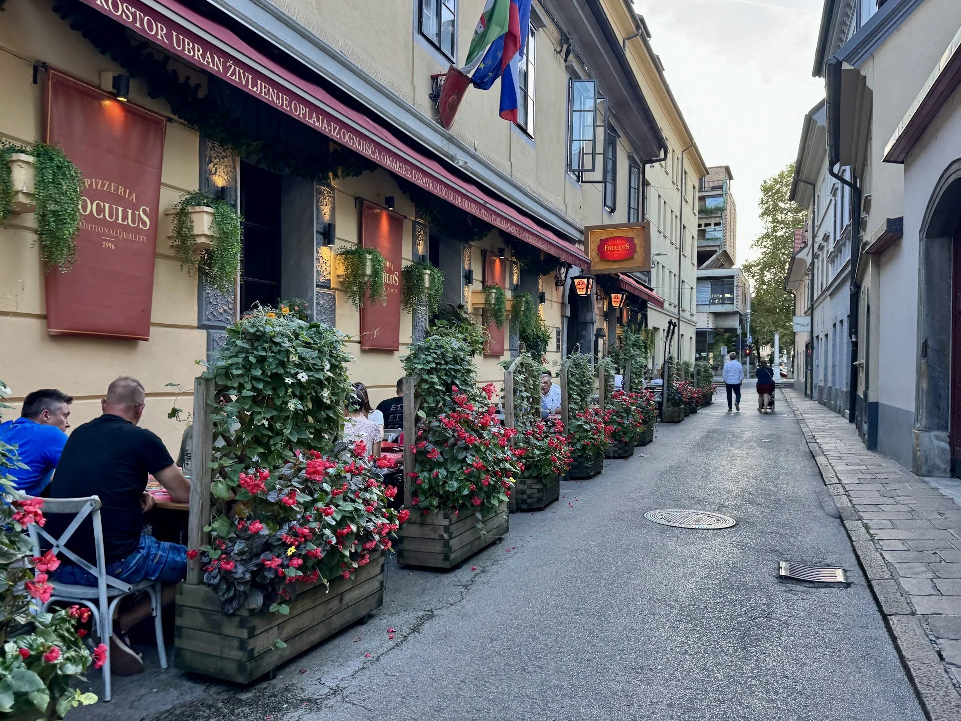 A European city sidewalk with outdoor seating, potted pink flowers, and a restaurant with a pink sign and striped red canopy. Pedestrians walk along the street, and buildings with open windows and flags are visible.