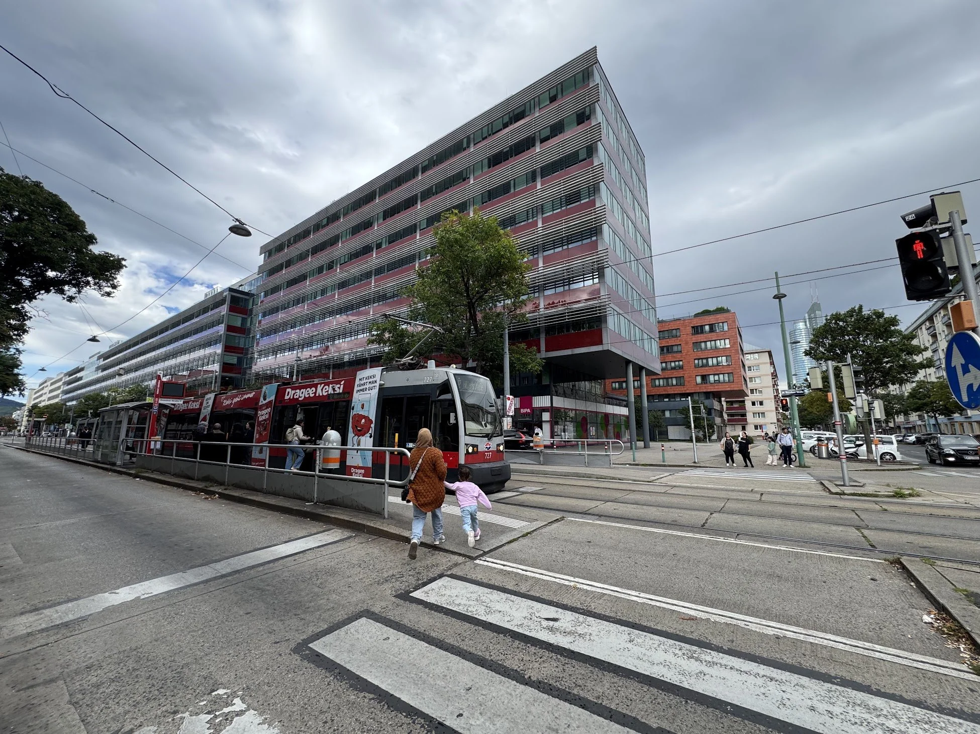 City street scene showing a modern multi-story office building, a tram, traffic lights, and pedestrians, including children, under a cloudy sky.