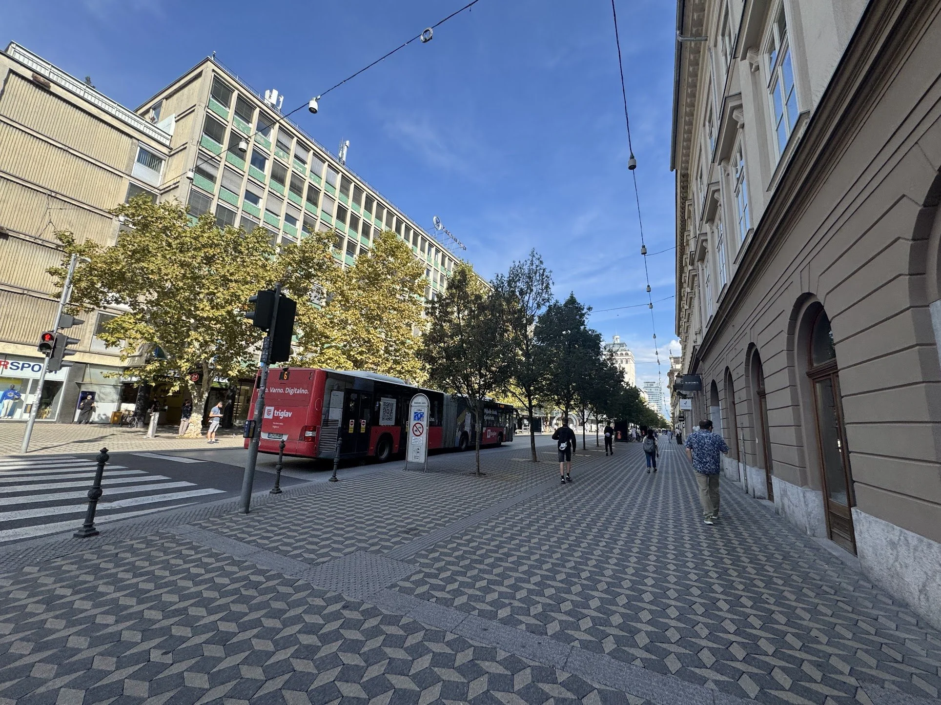 City street scene with a red bus, people walking on the sidewalk, a beige building on the right, a large modern building on the left, trees, crosswalk, and overhead wires against a blue sky.