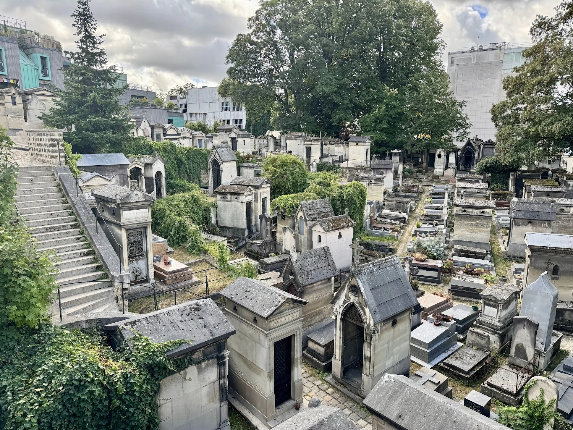 Aerial view of an old cemetery with numerous tombstones and mausoleums surrounded by greenery and tall modern buildings, with cloudy sky overhead.