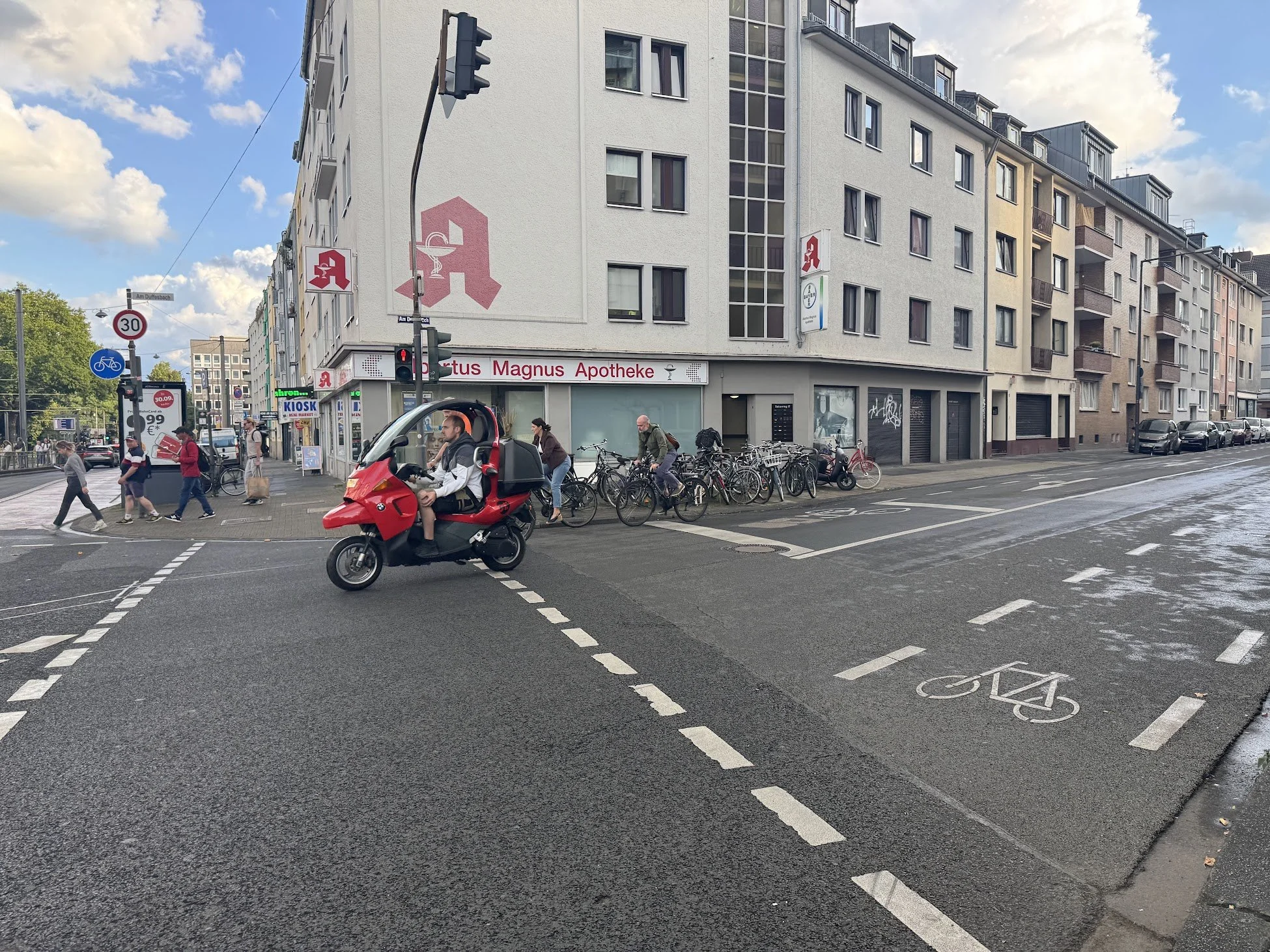 City street scene with a pharmacy named 'Fortus Magnus Apotheke' on a corner, pedestrians walking, bicycles parked along the sidewalk, a scooter on the road, traffic signals, and a building with windows and balconies, under a partly cloudy sky.