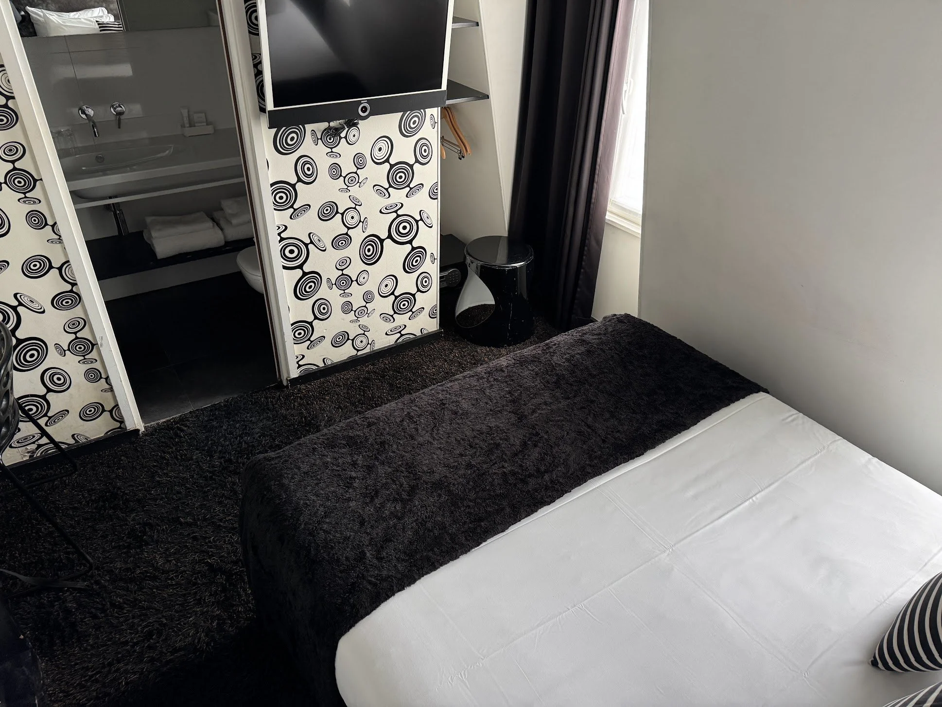 A hotel room with a bed, black fur throw, and white bedding. A black side table and a flat-screen TV mounted on the wall are visible. There is a small closet area with hangers, a bathroom with a sink and towels, and a window with dark curtains.