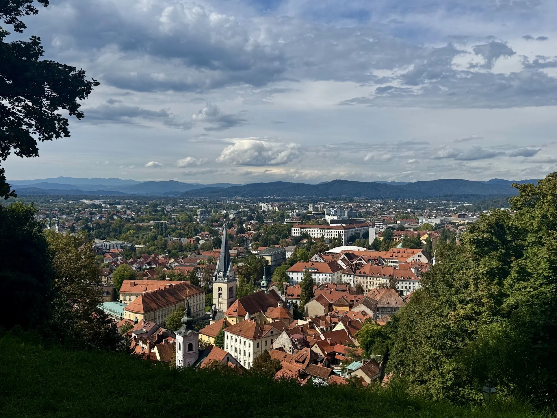 A panoramic view of a European town with red-tiled roofs, a church with a tall steeple, surrounded by green trees, with mountains and a cloudy sky in the background.