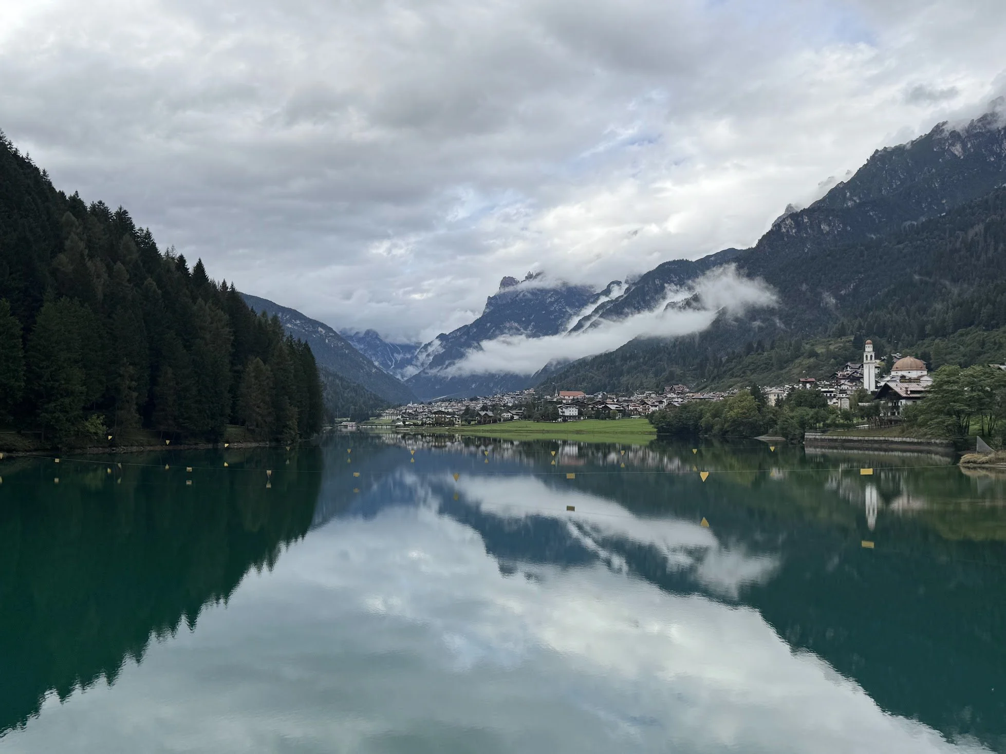 Scenic view of a mountain village with houses, church, and lush greenery, reflected in a calm lake, surrounded by towering mountains with cloud cover.