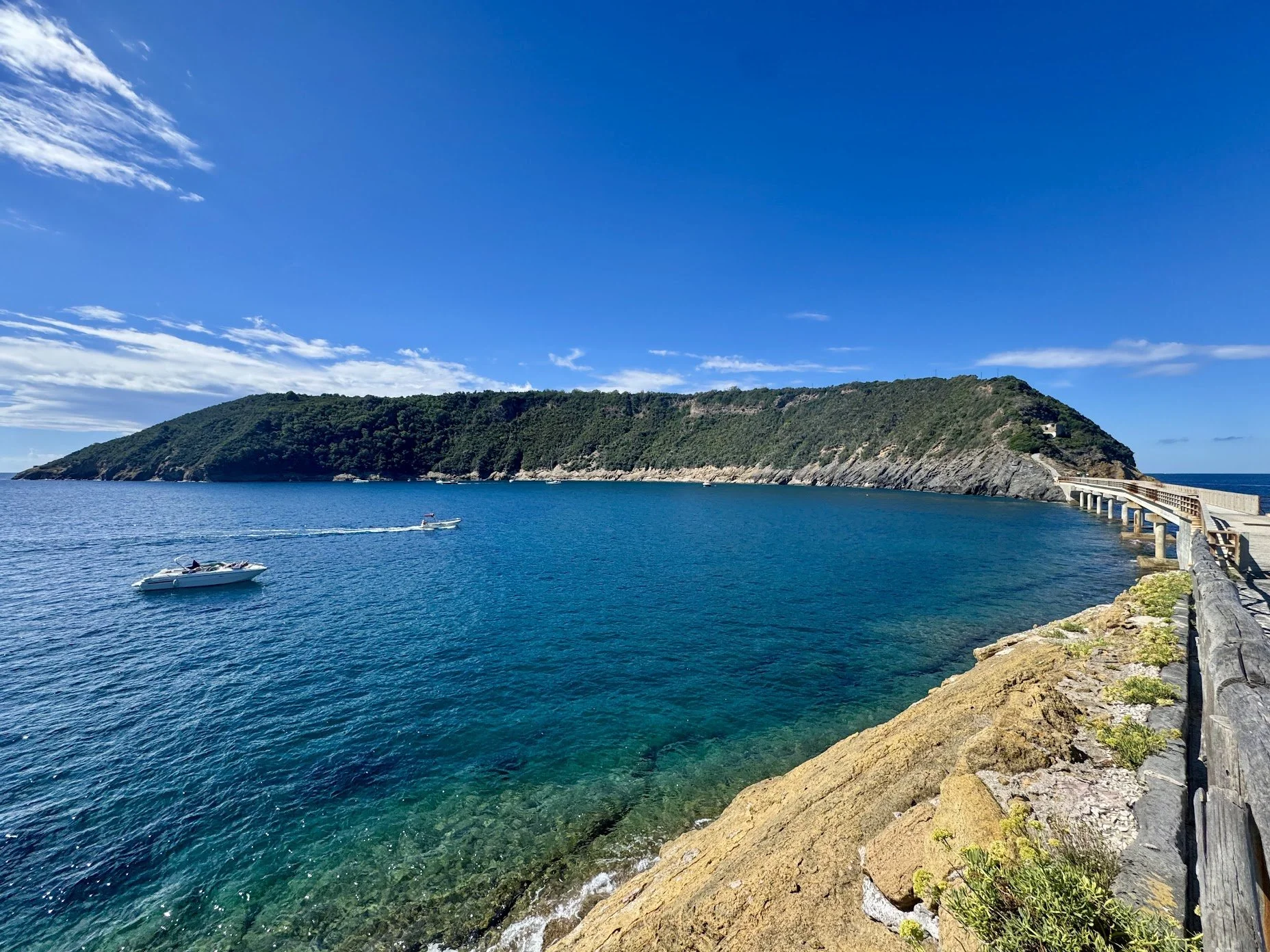A scenic view of a blue ocean with a white boat and a small yacht. In the background, there is a lush green hill with a pathway along the coastline and a bridge extending into the water under a bright blue sky with some white clouds.