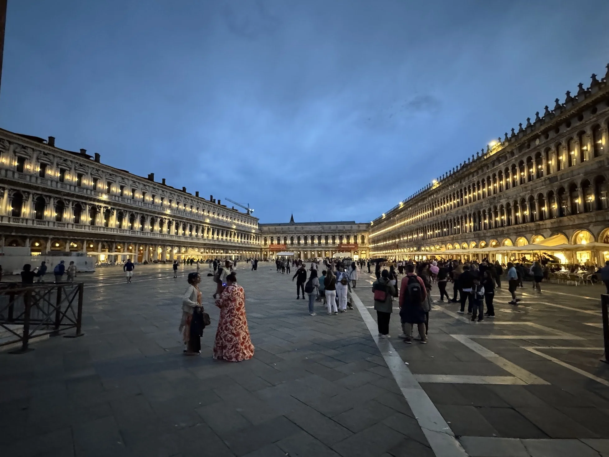 Venetian square with people walking and two large, illuminated classical buildings on either side, under a twilight sky.