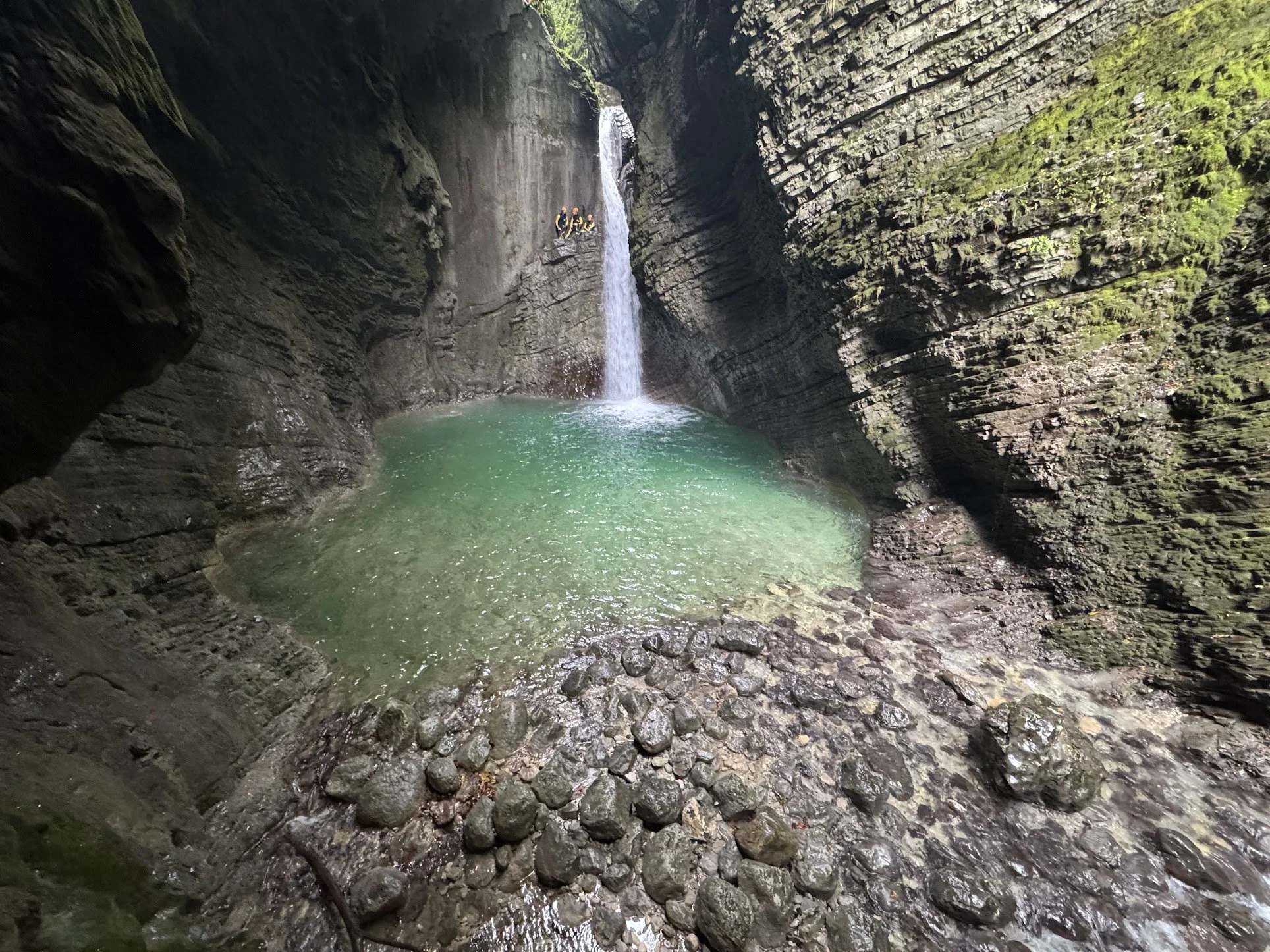 People sitting on rocks next to a waterfall in a canyon with steep rocky walls covered in moss and greenery, with clear water at the bottom.