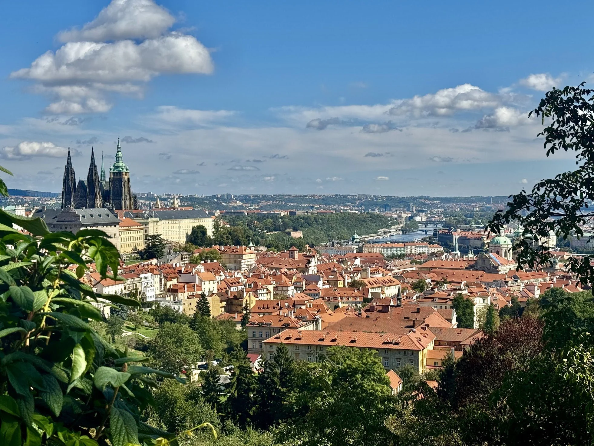 A panoramic view of Prague cityscape with historic buildings, red rooftops, a river, and a cathedral with two tall spires, under a blue sky with scattered clouds.