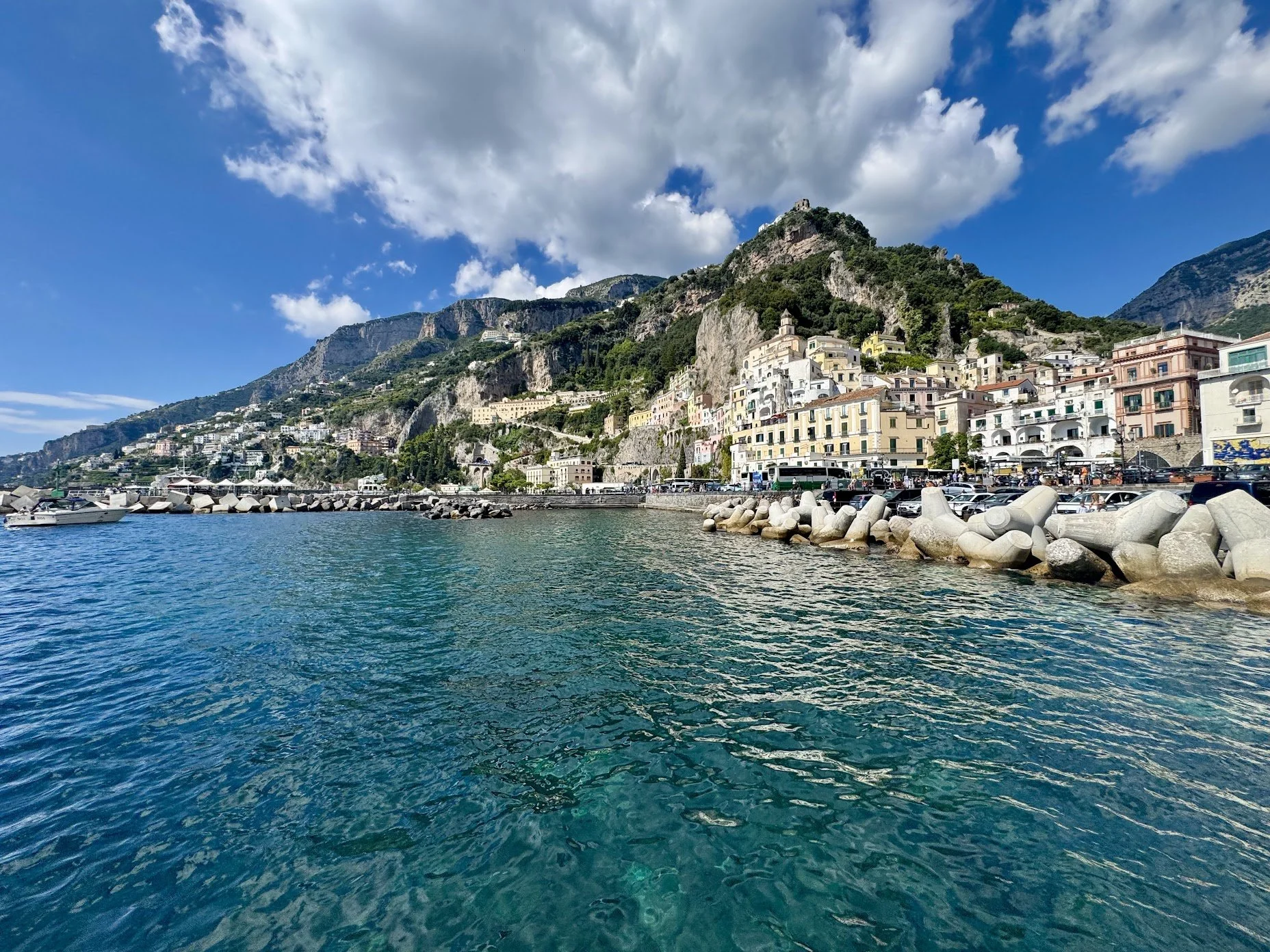 Colorful buildings on a hillside above a harbor with boats, stone breakwater, and rocky shoreline under a partly cloudy blue sky.