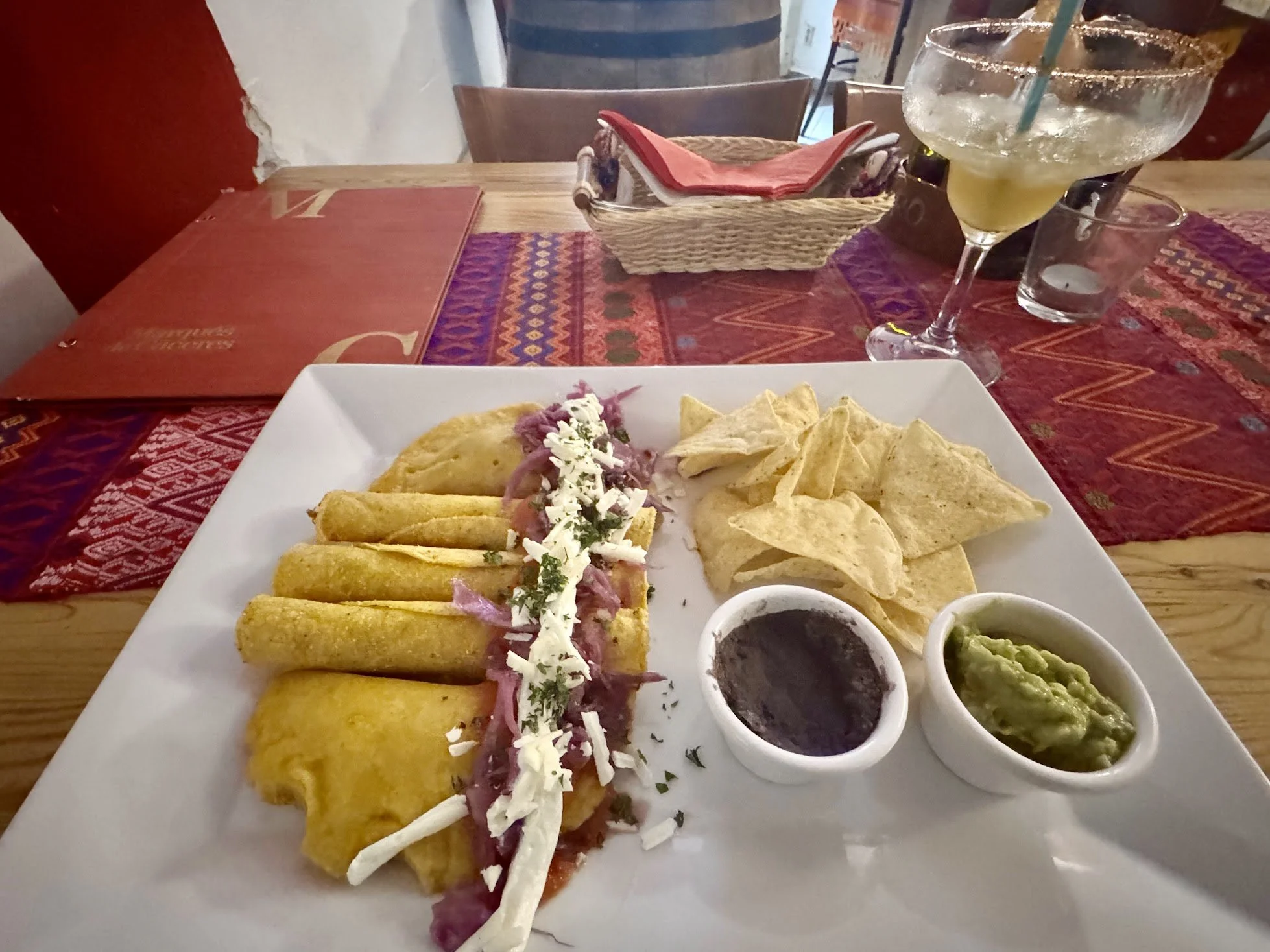 Plate of crispy tacos topped with shredded cheese and pico de gallo, with side dips of guacamole, salsa, and sour cream. In the background, a margarita with a salted rim, lime slice, and blue straw, along with a glass of water, on a red patterned tablecloth at a restaurant.