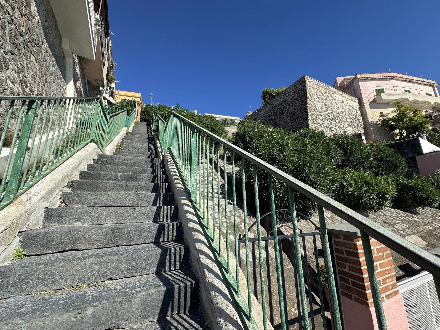 Stone staircase with green metal railings leading uphill, flanked by buildings and greenery, against a bright blue sky.