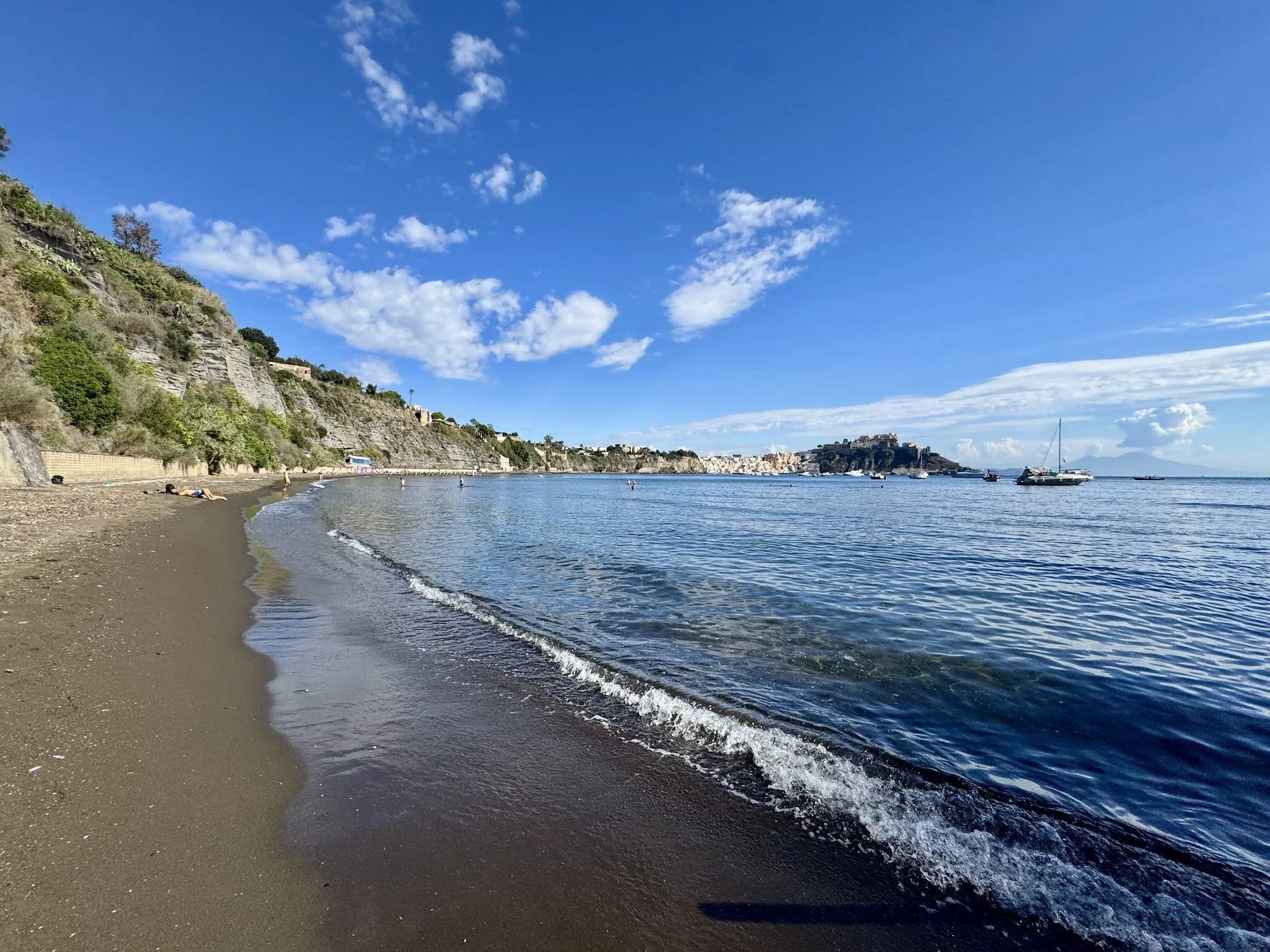 A coastal scene with a sandy beach, gentle waves, and a hillside with houses and greenery. Several boats are anchored in the water, and the sky is bright blue with some scattered clouds.