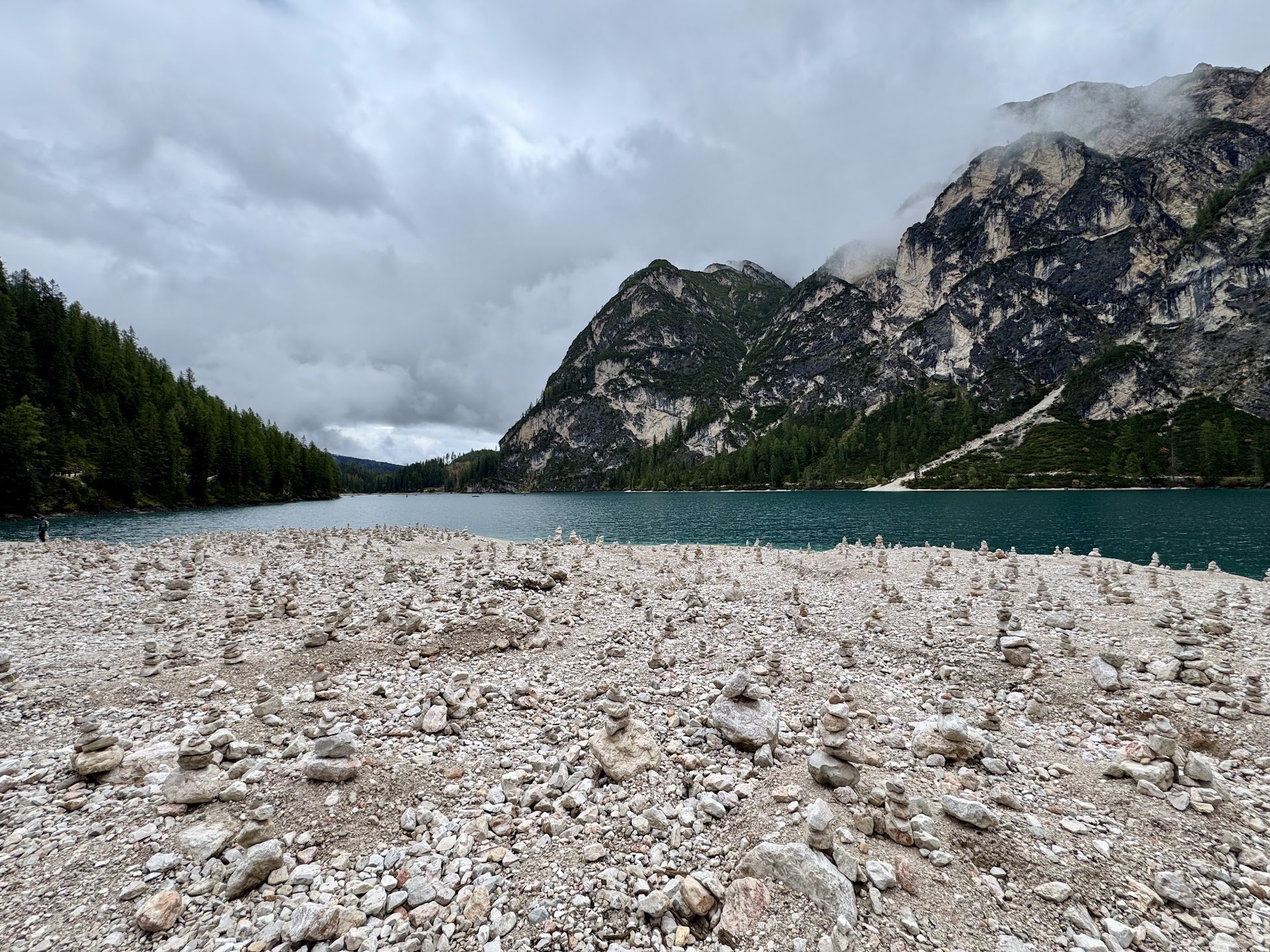 Rocky shoreline with large stones in the foreground, a body of water, green forested mountains, and a cloudy sky in the background.