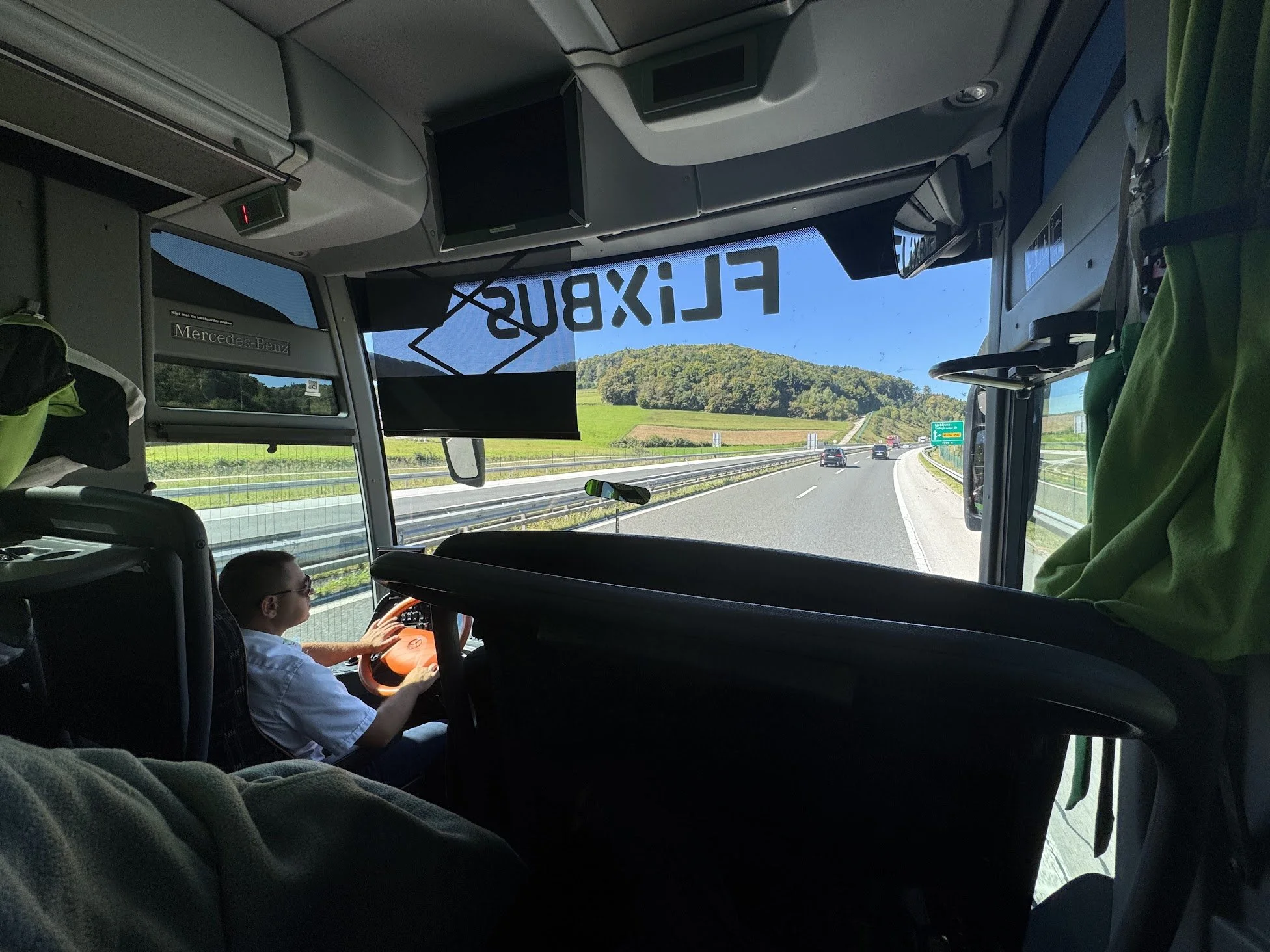 Inside a bus traveling on a highway with green hills visible outside. A person in a white shirt is sitting in a child seat, holding the steering wheel, and looking ahead. Overhead monitors, signage, and seats are visible inside the bus.