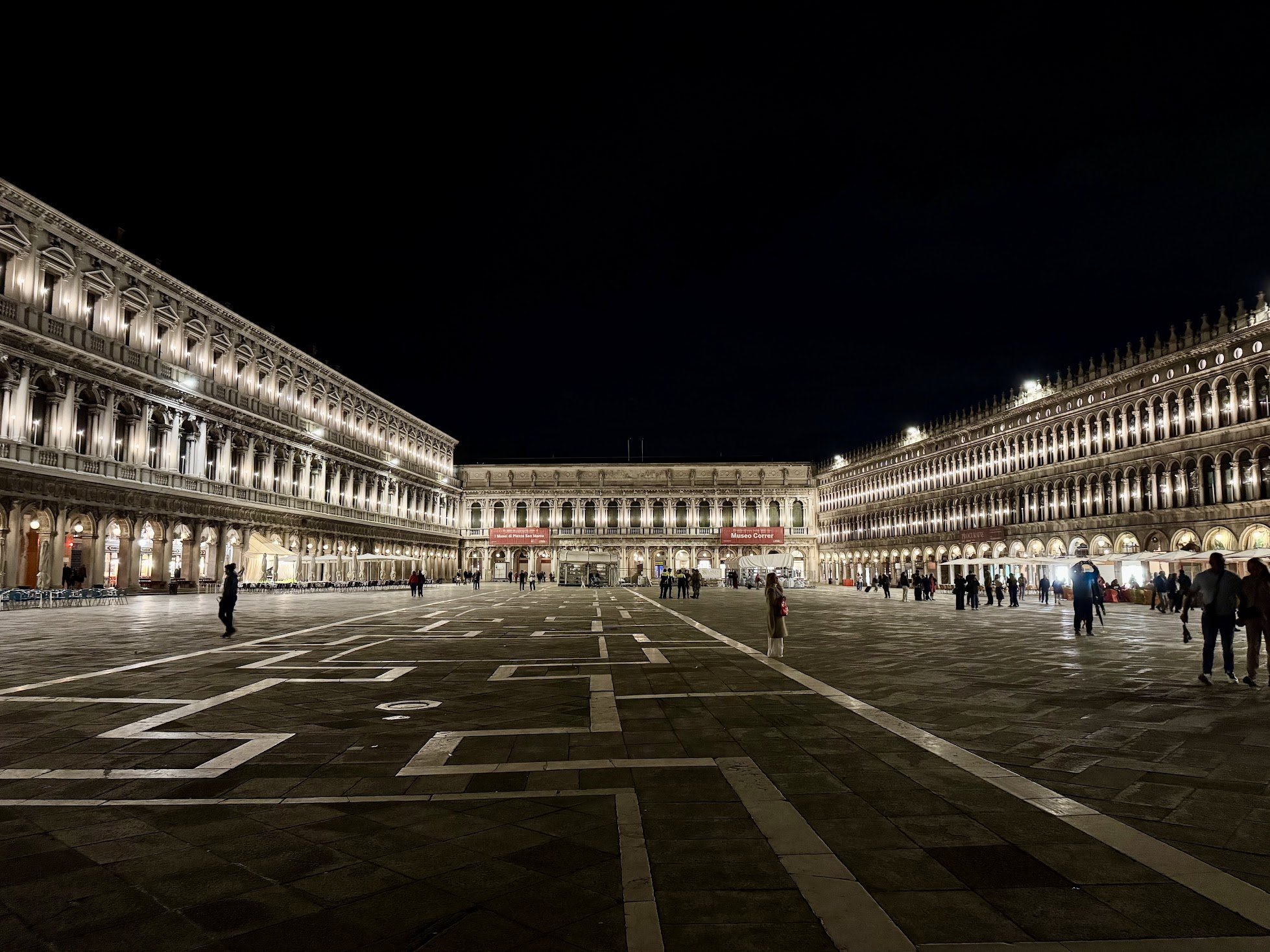 Night view of a large open square surrounded by illuminated historic buildings with arched windows, with a few people walking and standing around.