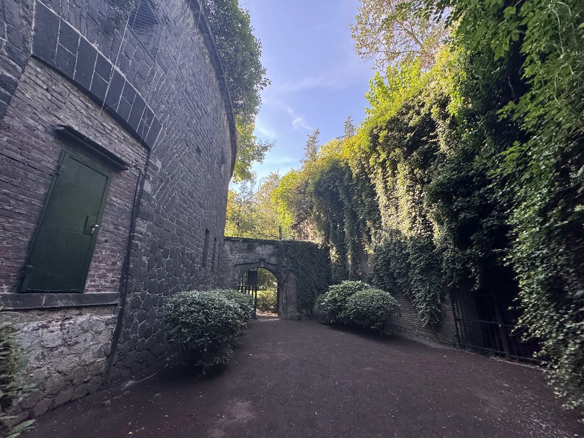 A stone wall with a green door on the left and lush green trees lining a pathway under a clear blue sky.