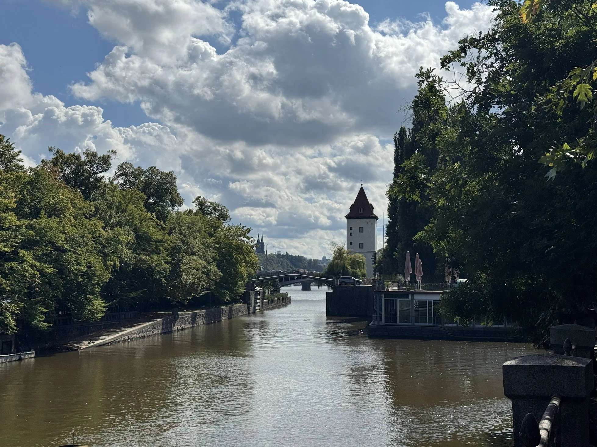 A river runs through a city with trees on both sides. A tower with a pointed roof is visible in the distance, with clouds scattered across a blue sky.