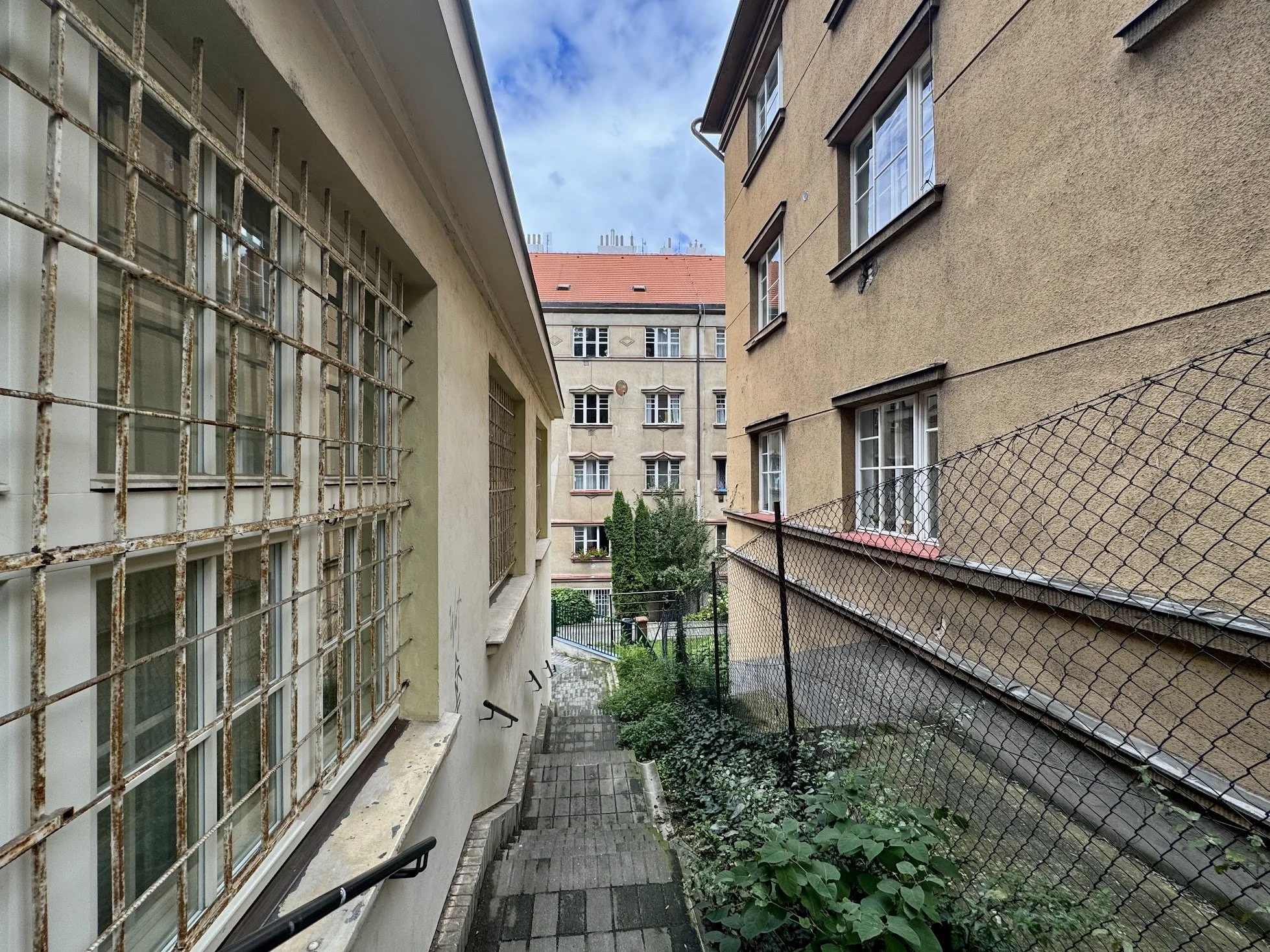 View of a narrow outdoor pathway between two residential buildings, with a rusty metal window grill on the left and a chain-link fence on the right, leading to a green backyard with trees. The sky is partly cloudy.