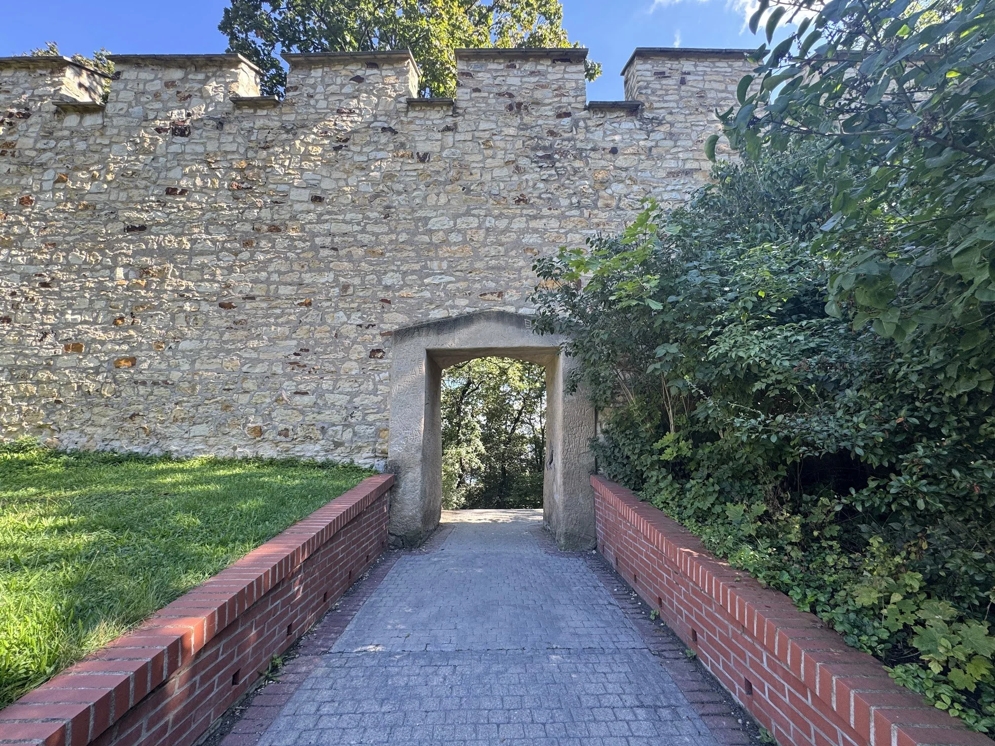 Stone fortress wall with an archway and greenery on the right side, brick-lined pathway leading through the arch, blue sky in the background.