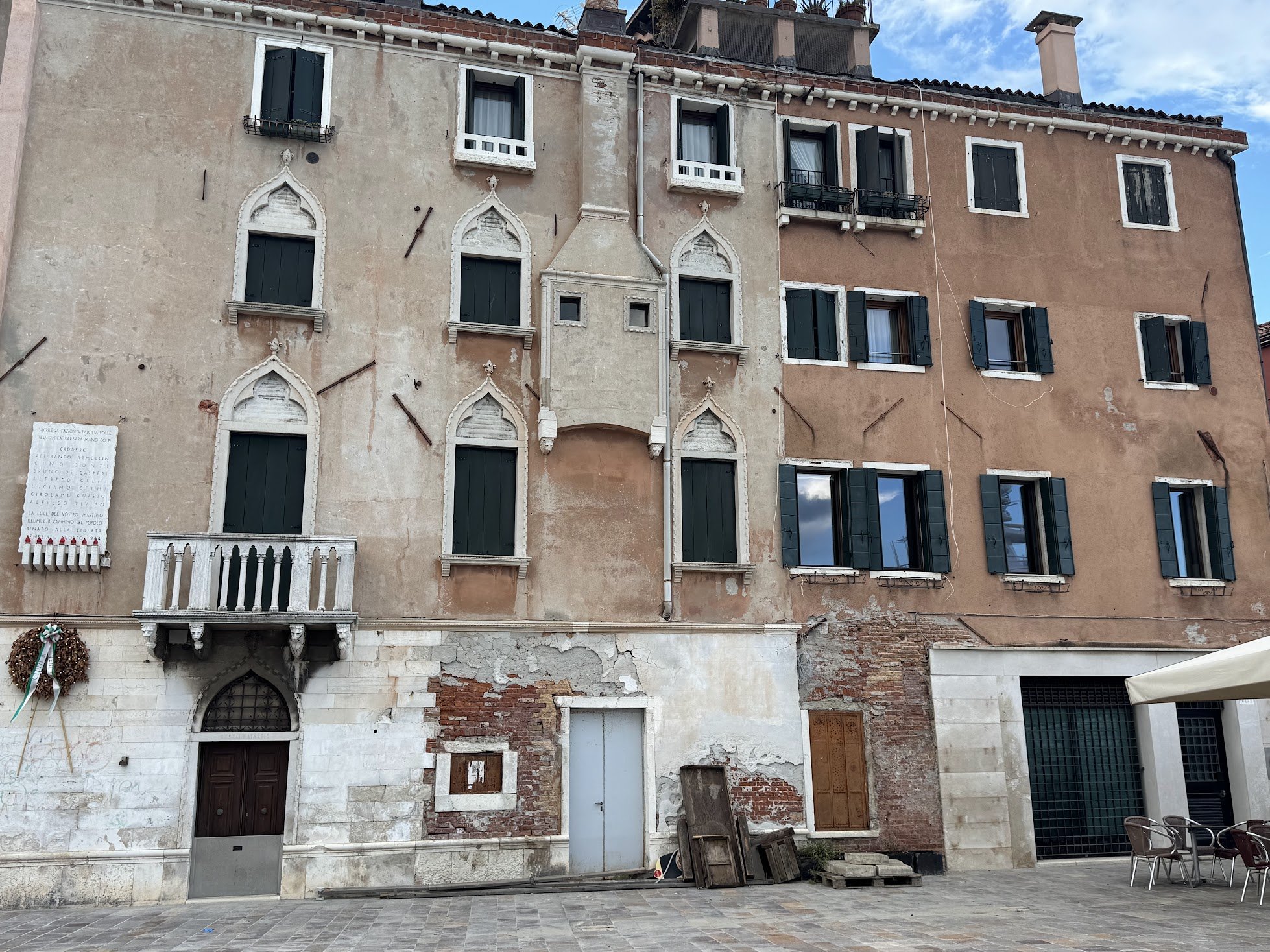 Historic building with damaged plaster and exposed brick, several boarded-up windows, and a small balcony. There are chairs on the right side and some wooden pallets and old doors at the bottom. The sky is partly cloudy.