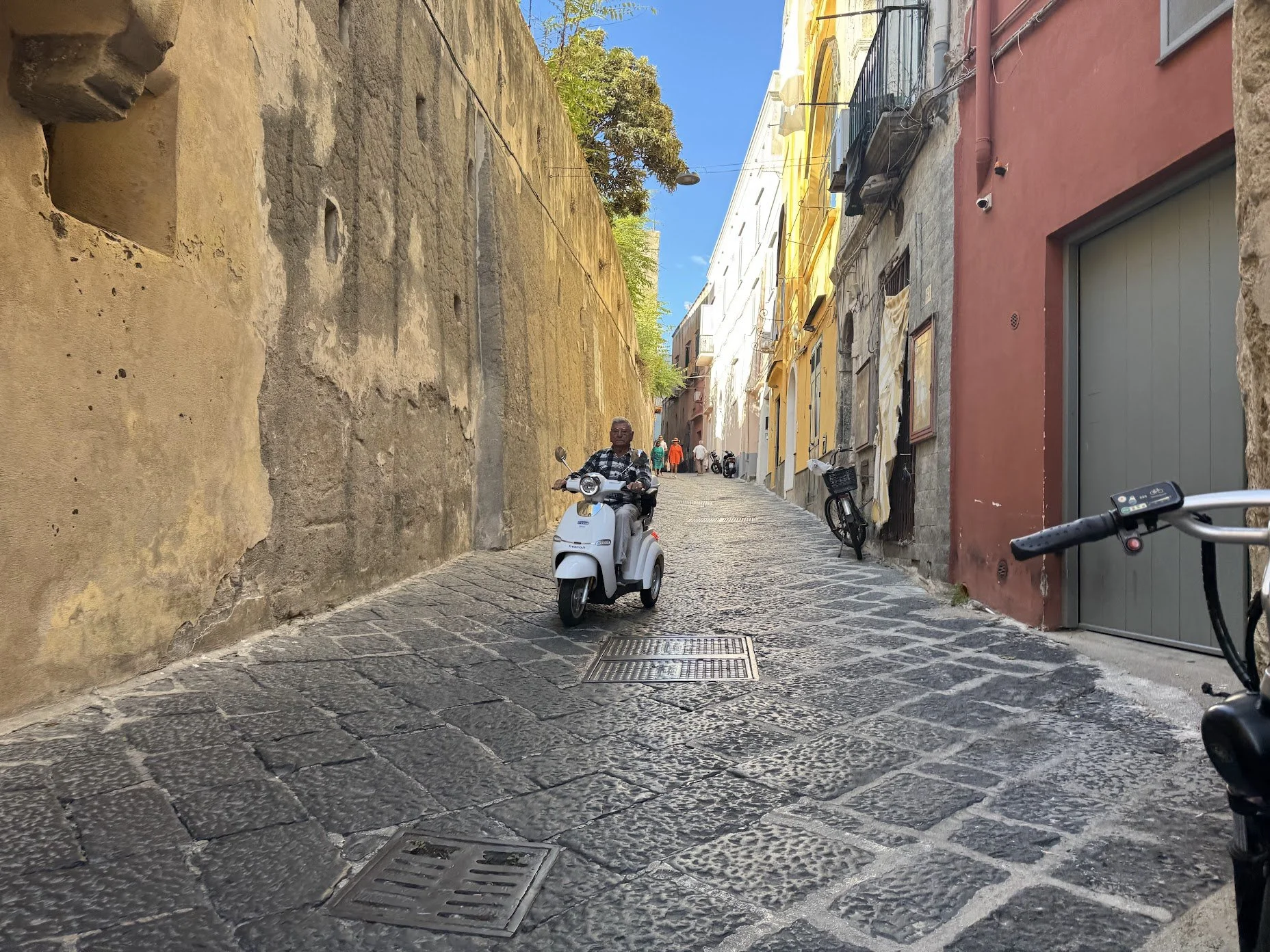A narrow cobblestone street with a man riding a white scooter, tall colorful buildings on the right, a large stone wall on the left, and a few pedestrians walking in the background. The sky is clear and blue.
