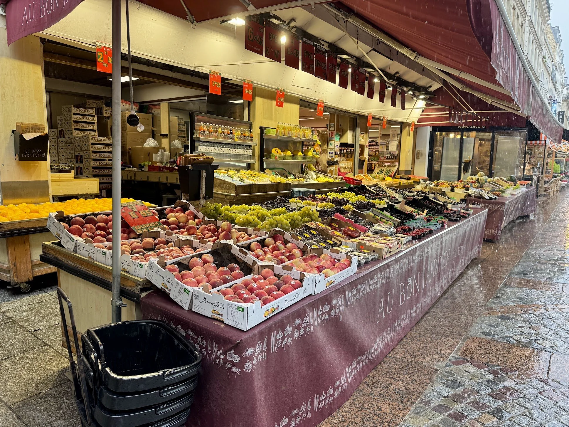 A street market stand under rain, displaying various fresh fruits such as peaches, grapes, and melons. Rain is visible on the wet pavement and a glass enclosure is seen in the background.