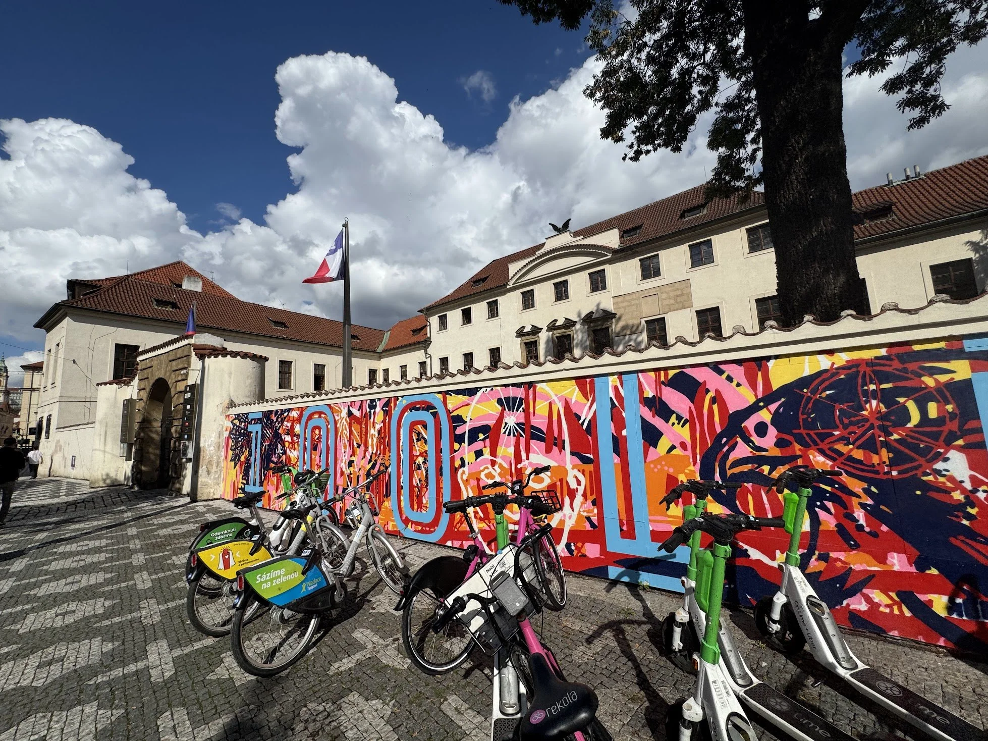 A row of parked rental bikes in front of a colorful mural on a city street. Behind the bikes, there is a historic building with a red tiled roof, windows, and a flagpole with a flag, under a partly cloudy sky with a large tree.