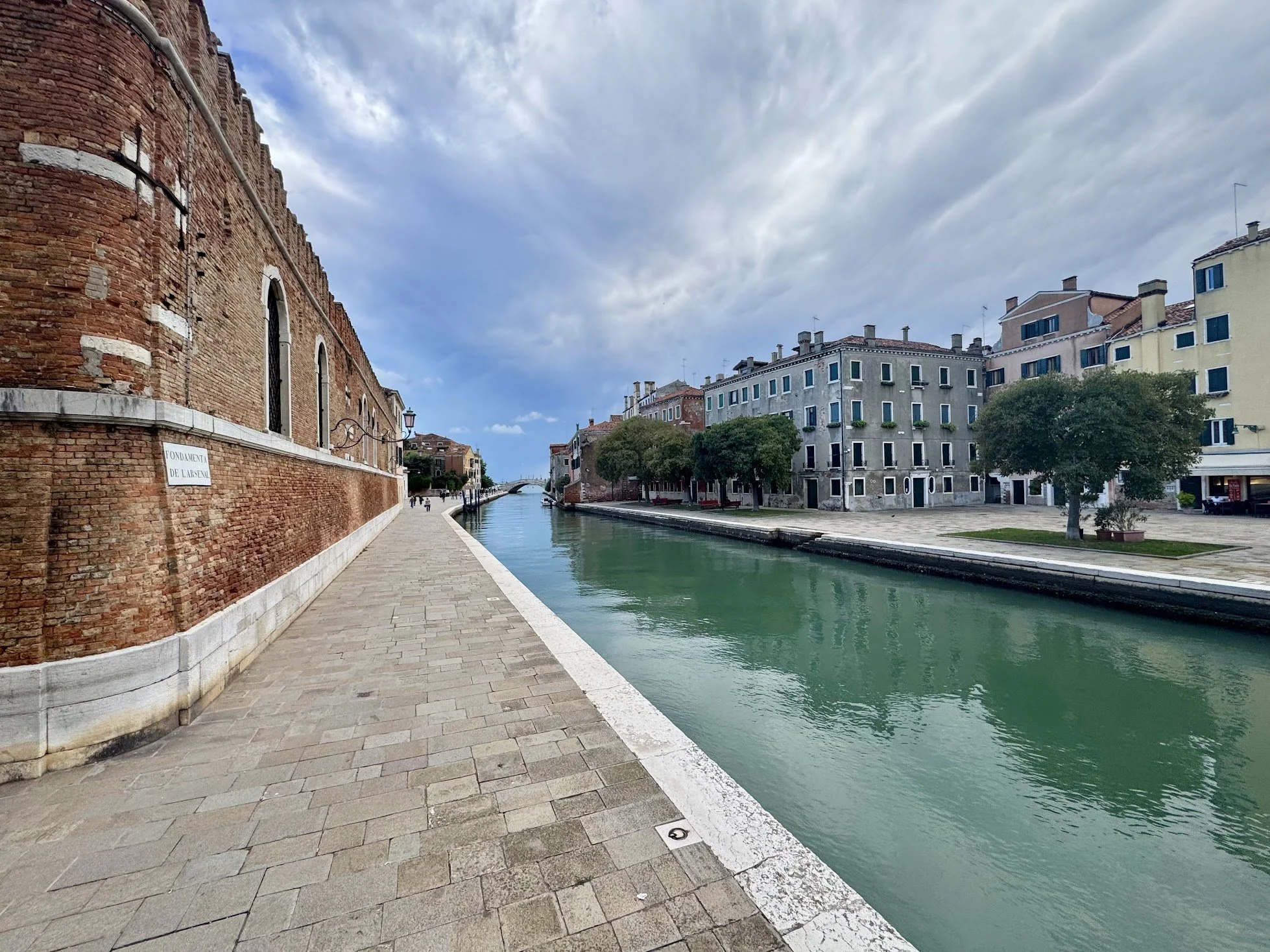 A scenic view of a canal with a paved walkway along it, surrounded by old brick and modern buildings under a partly cloudy sky.