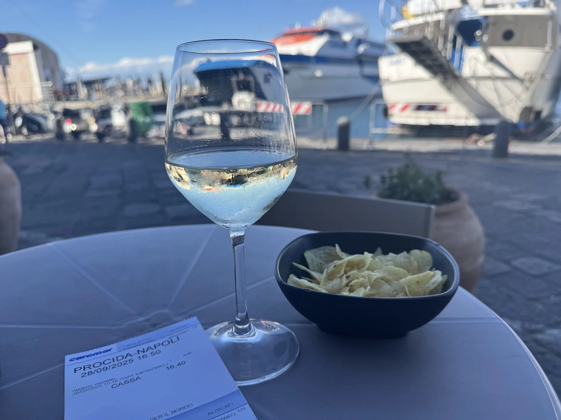 A glass of white wine, a bowl of pasta, and a restaurant receipt on a table at a marina with boats in the background.