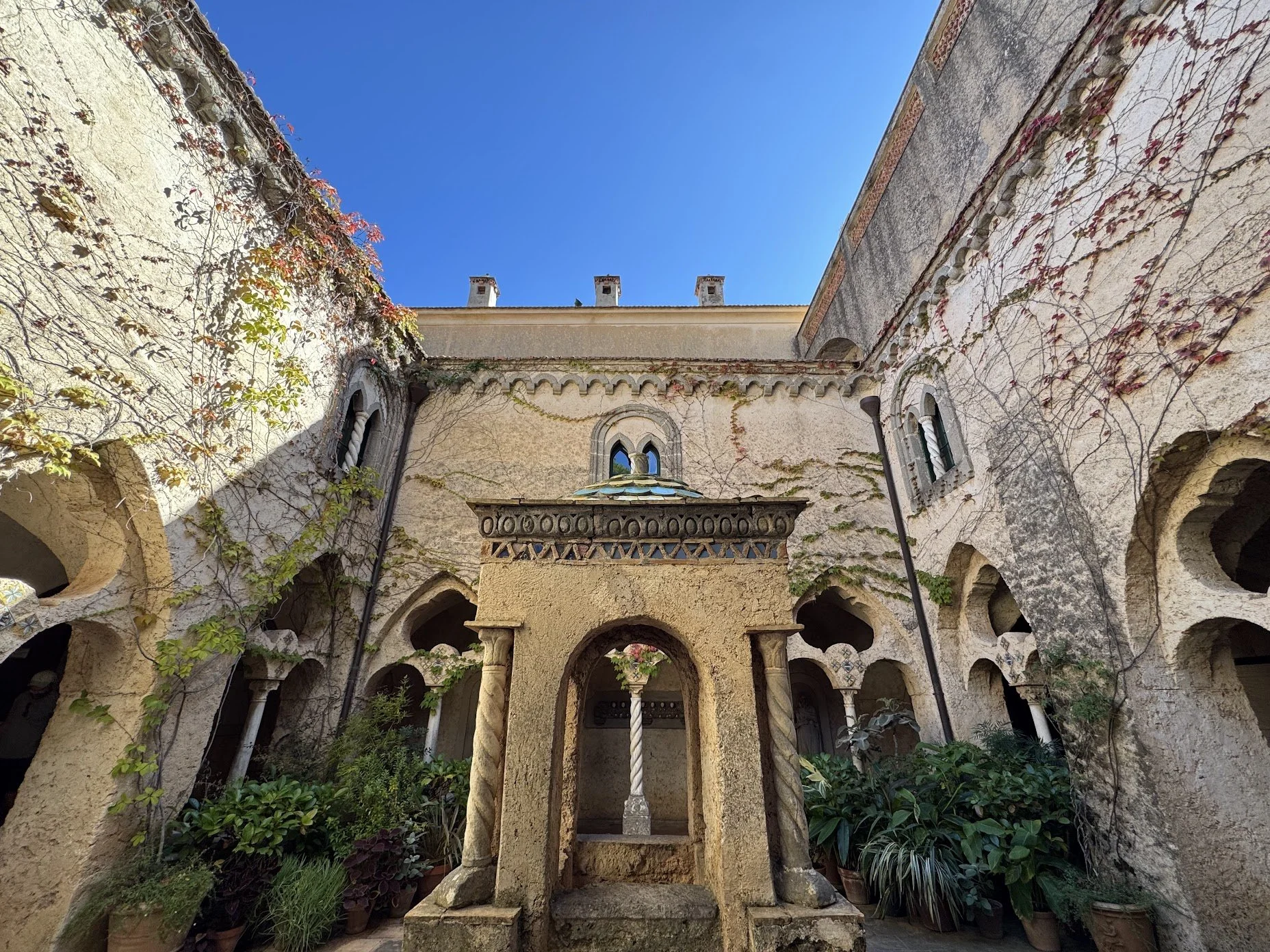 Courtyard view of an old stone building with arched windows and a central stone fountain, surrounded by potted plants and vines climbing the walls, under a clear blue sky.