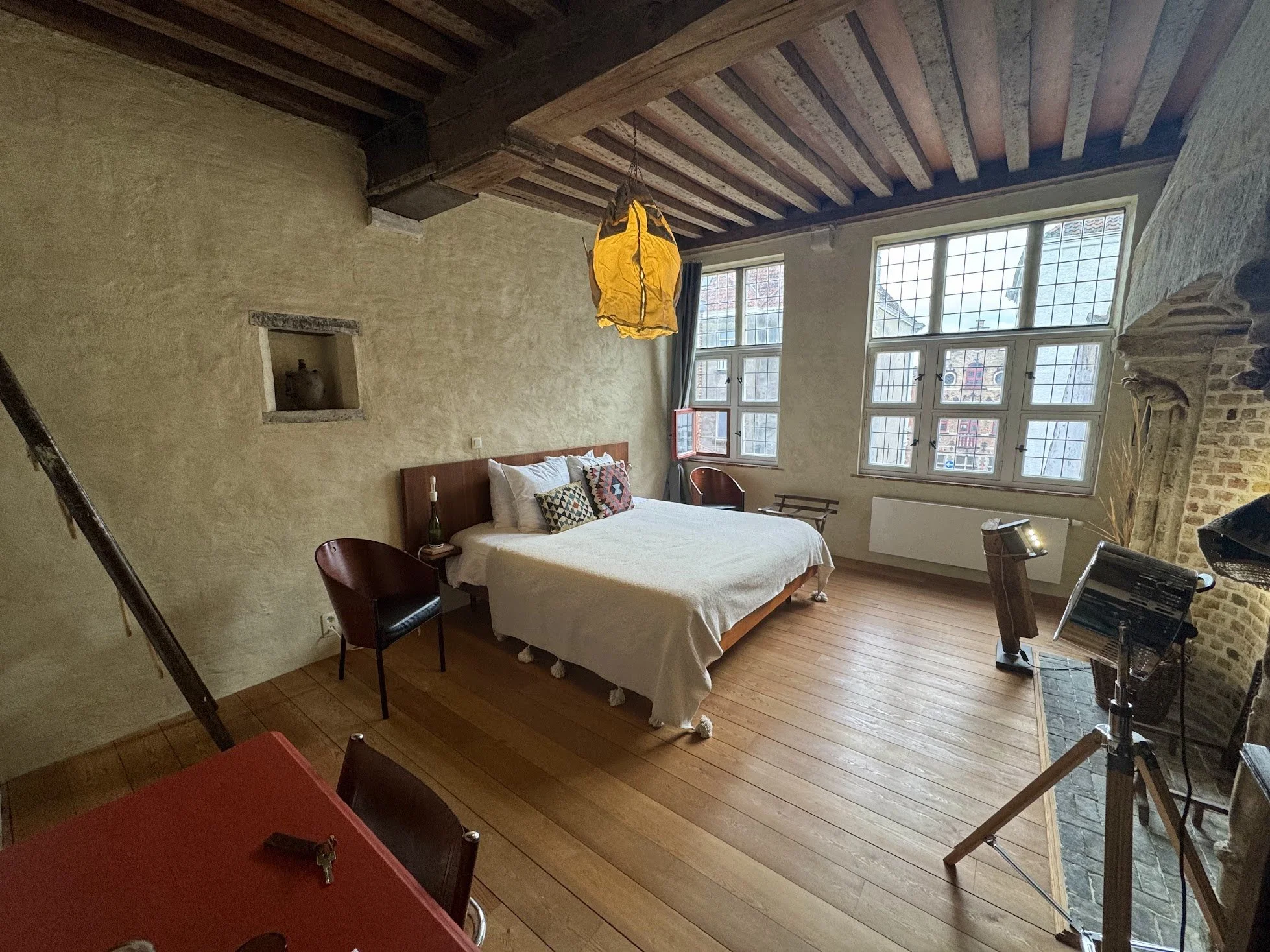 A cozy bedroom with a large bed, wooden floor, and windows letting in natural light. The room has a rustic feel with a wooden ceiling, brick accents, and a yellow pendant light hanging from the ceiling. There are some chairs around and a fireplace on