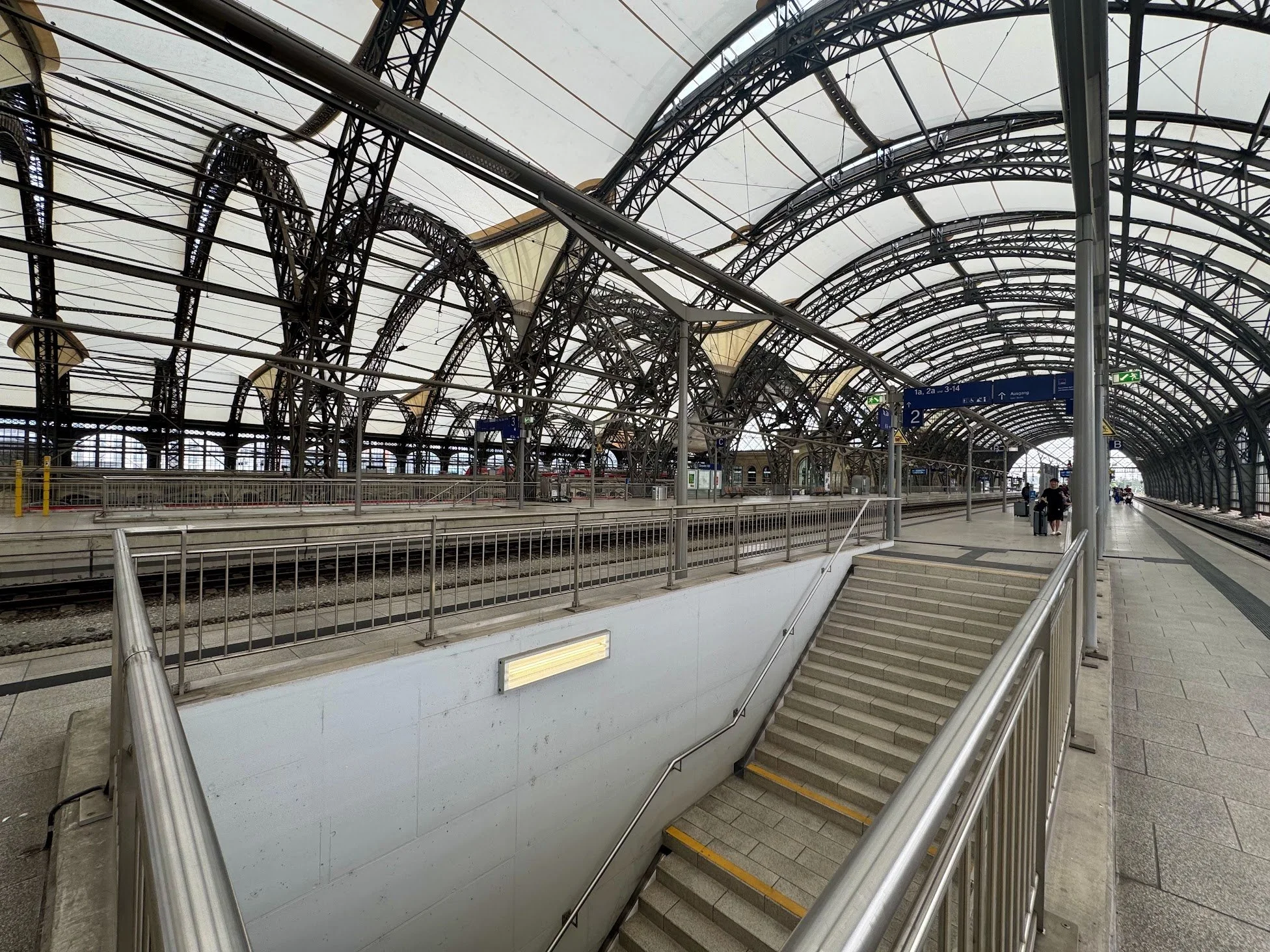 Interior of a train station with arched glass and metal roof, train platform with some passengers, signs indicating platform numbers, and a staircase leading down to an underground area.