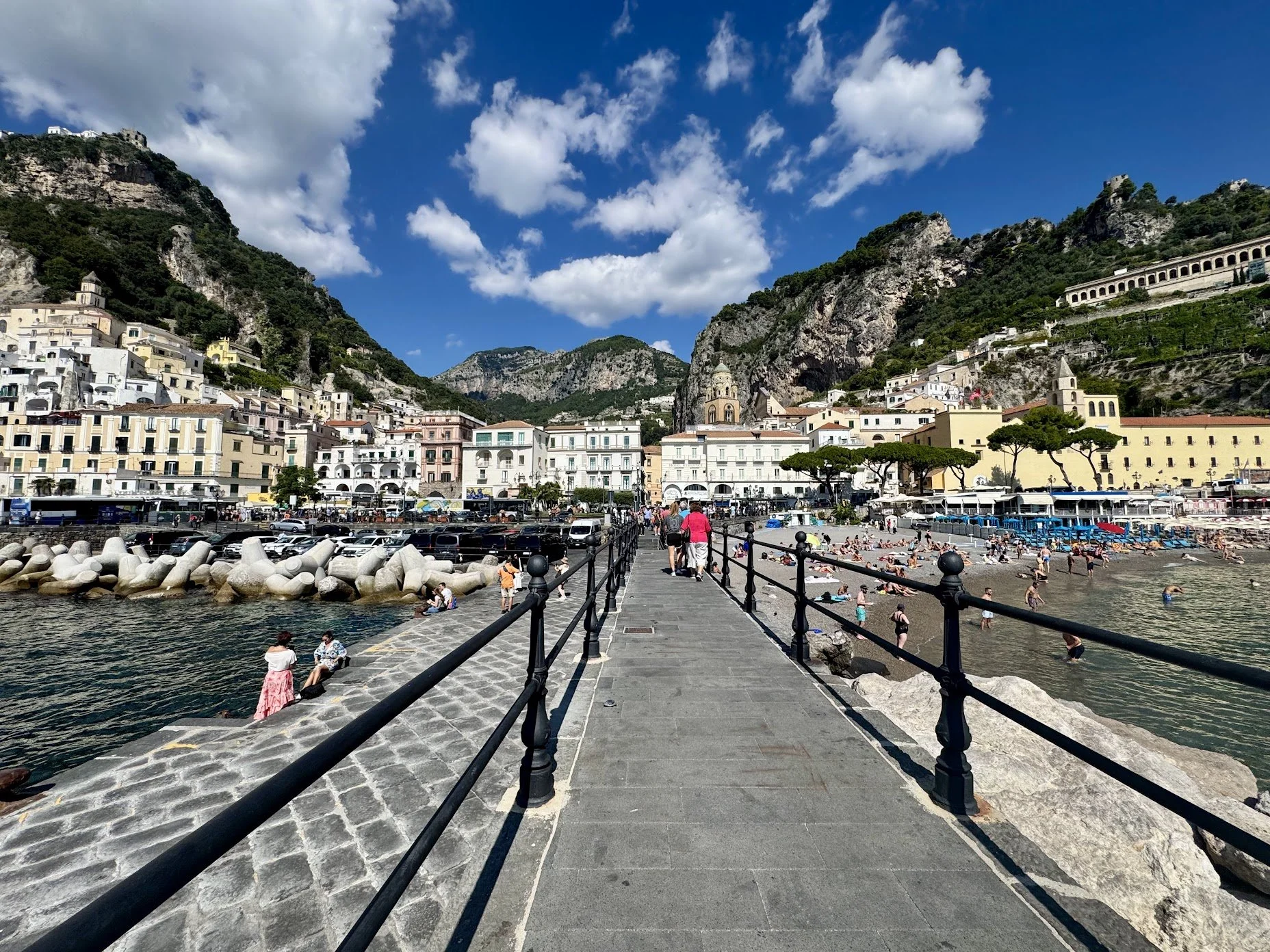 People walking on a stone pier along the coast, with beachgoers sunbathing and swimming, colorful buildings on hillside, hills and blue sky with clouds in the background.