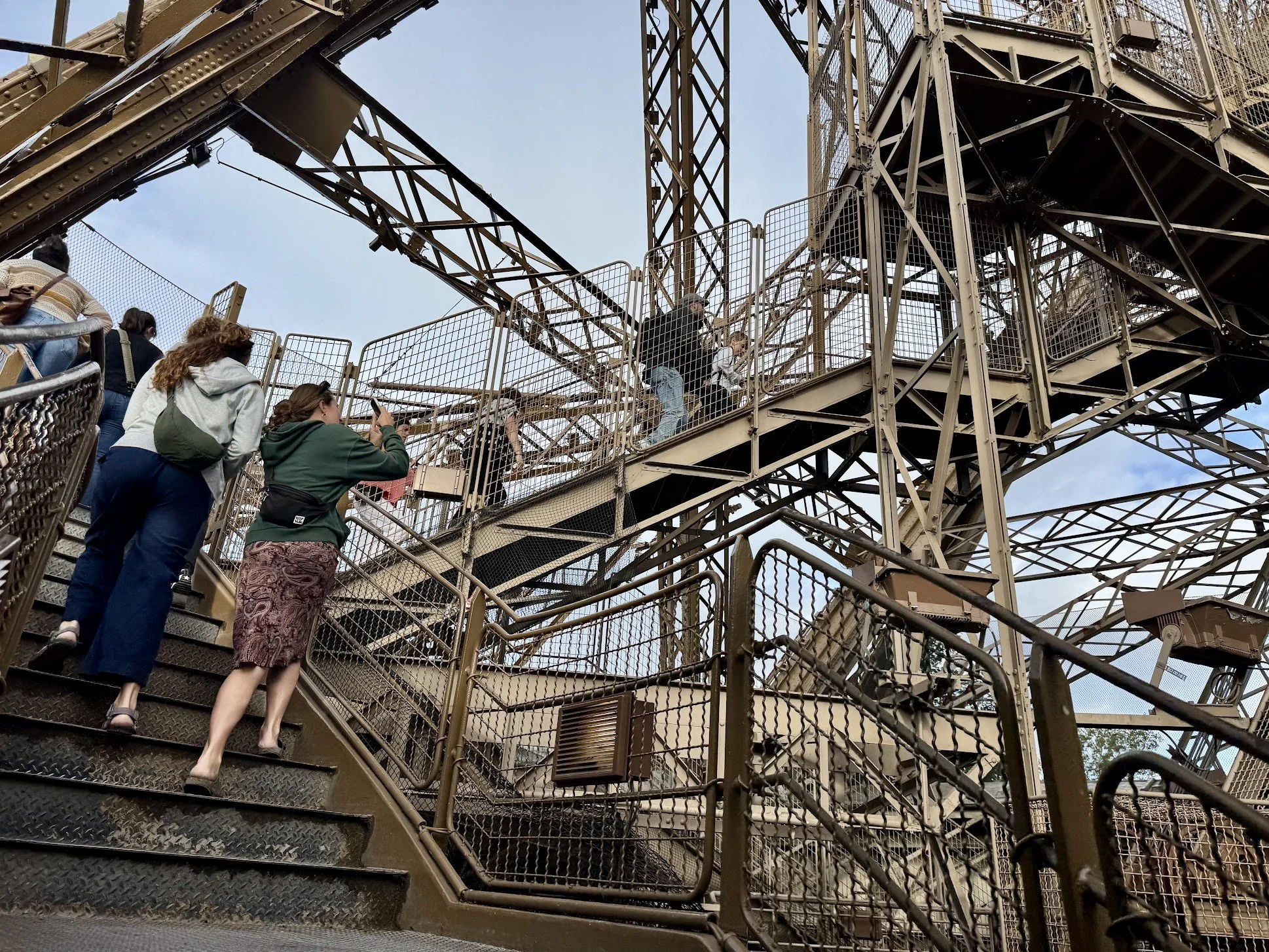 People walking up stairs on a large, intricate metal structure, possibly part of a Ferris wheel, with a partly cloudy sky in the background.