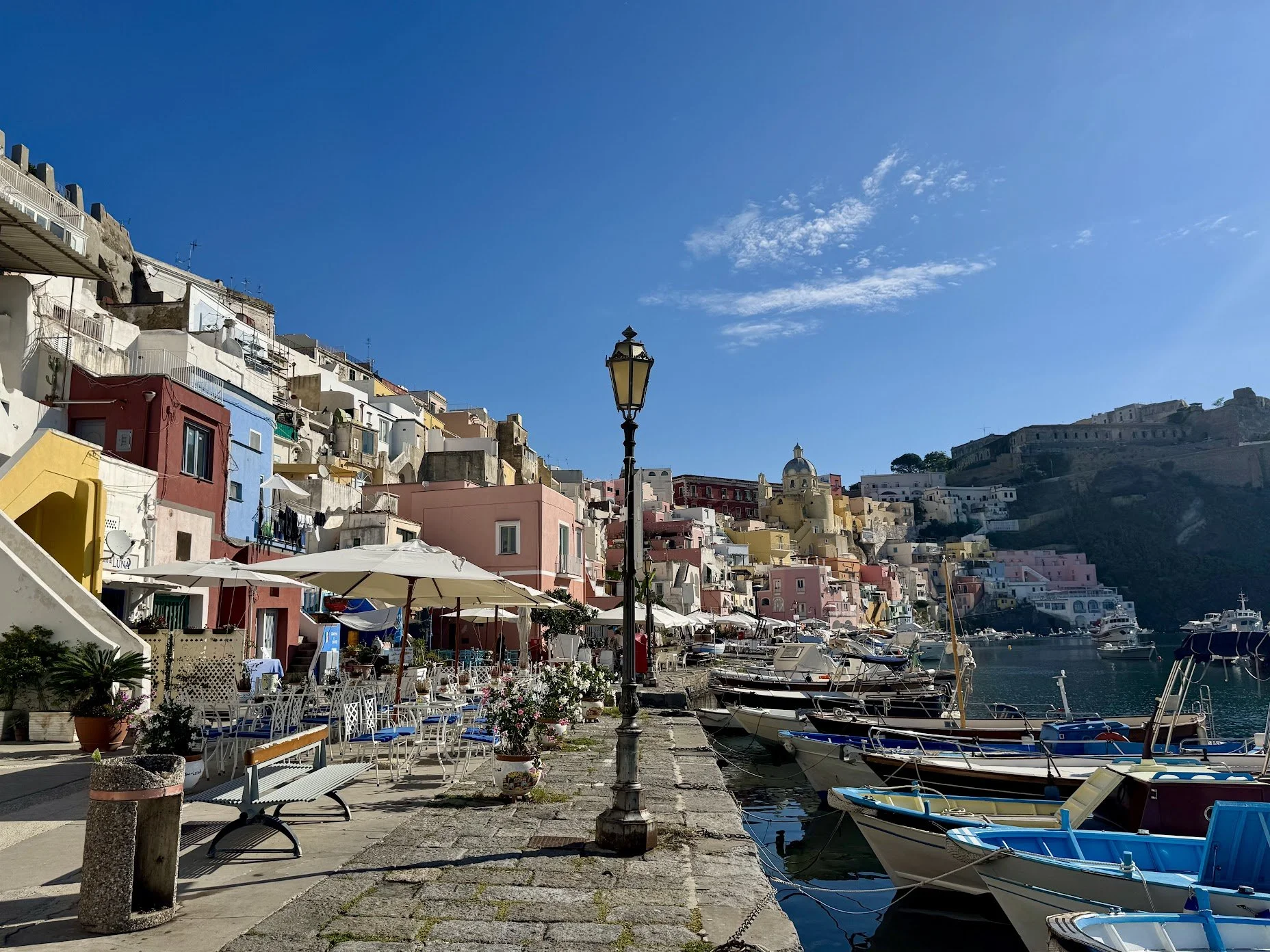 Colorful buildings on a hillside in a harbor town, boats docked along the waterfront, outdoor seating and a lamppost near the water, sunny day with a blue sky and some clouds.