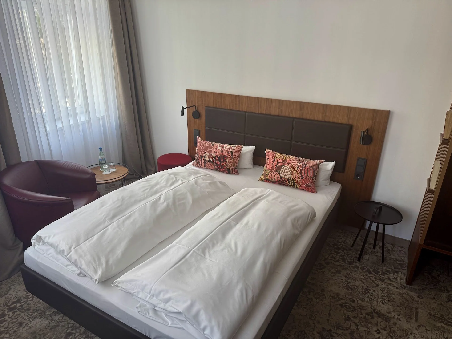 A hotel room with a double bed, colorful pillows, a wooden headboard, and a window with sheer curtains. There is a purple armchair, a small round table with a water bottle and a glass, and side tables with wall-mounted lamps.