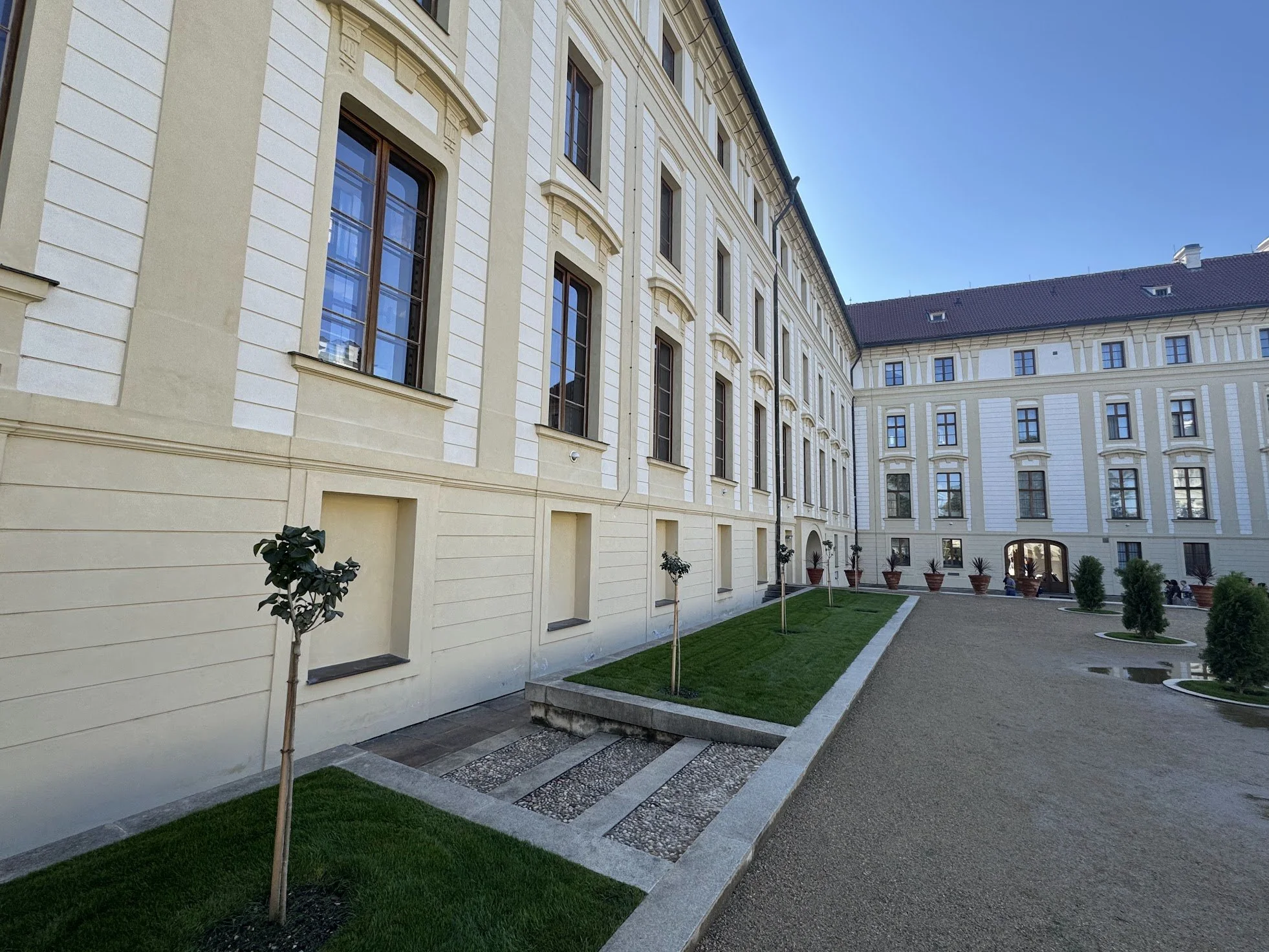 Corner of a large cream-colored building with multiple windows, small trees planted along the sidewalk, a well-maintained lawn, and a gravel pathway, under a clear blue sky.