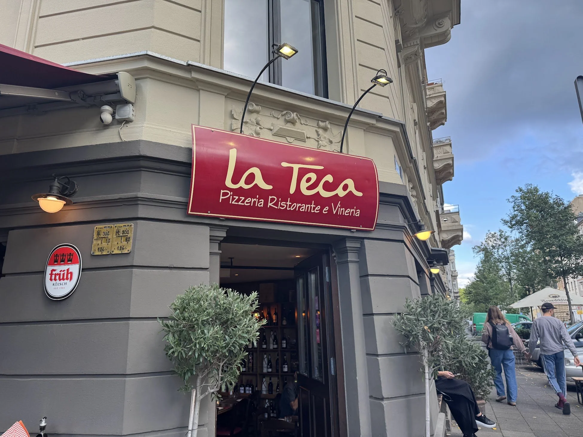 The storefront of 'la Teca,' an Italian restaurant and pizzeria, on a city street with people walking by. The sign is red with white text, and the building features decorative architectural details. There are outdoor street lights and greenery near t