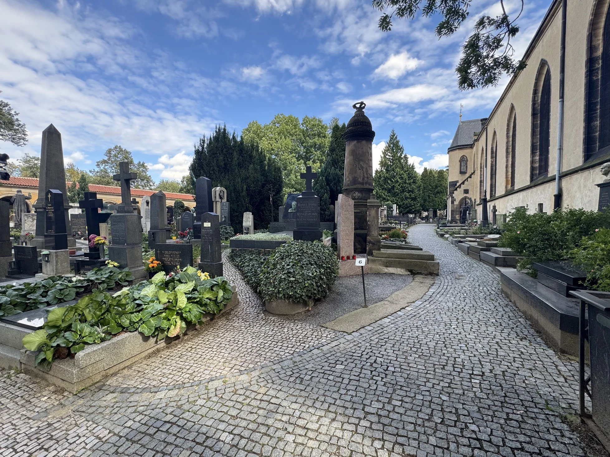 A cemetery with numerous tombstones and grave markers surrounded by trees, with a cobblestone pathway and a church building on the right side under a partly cloudy blue sky.