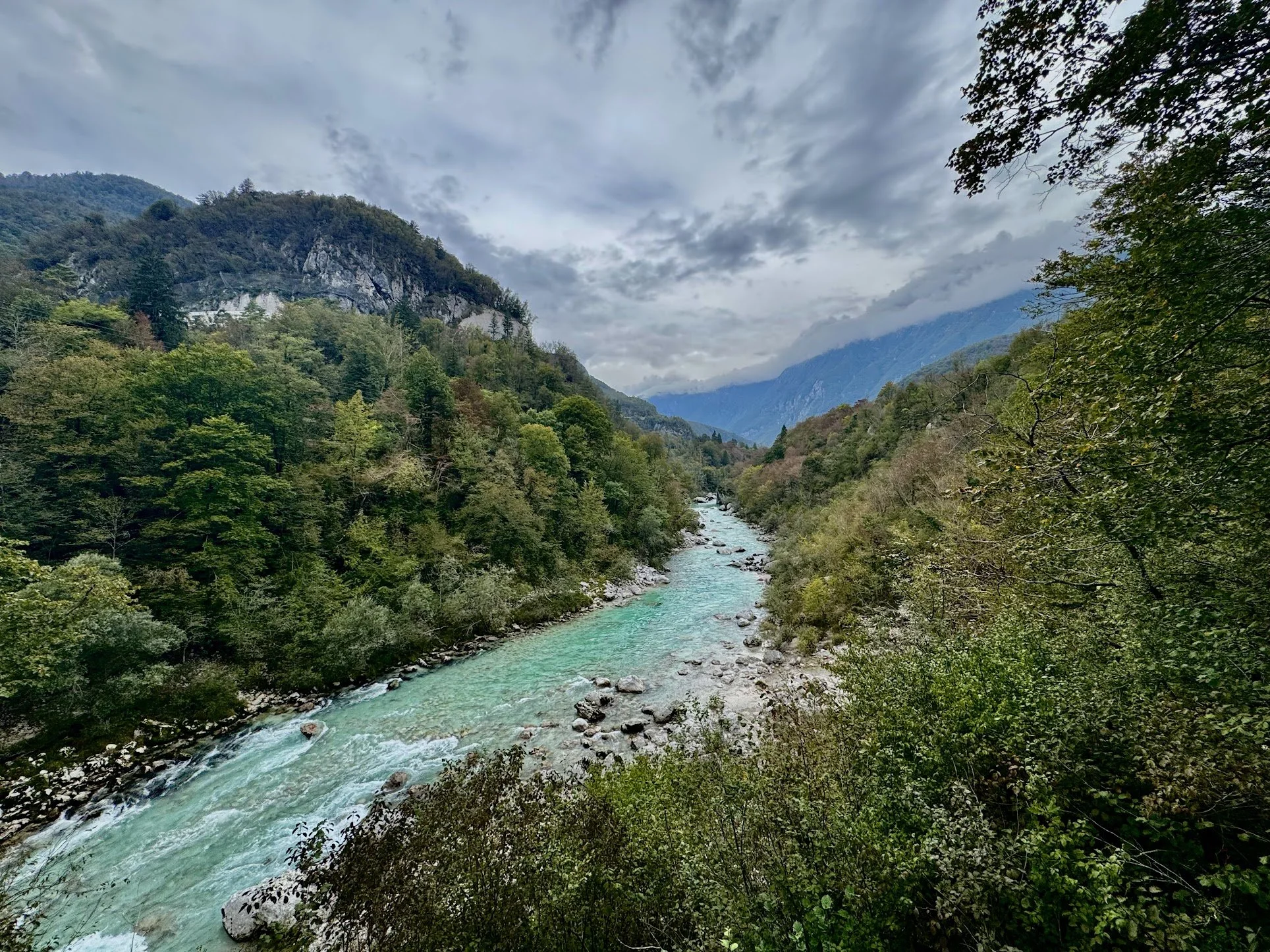 A scenic view of a river flowing through a lush green valley with forested mountains in the background under a cloudy sky.