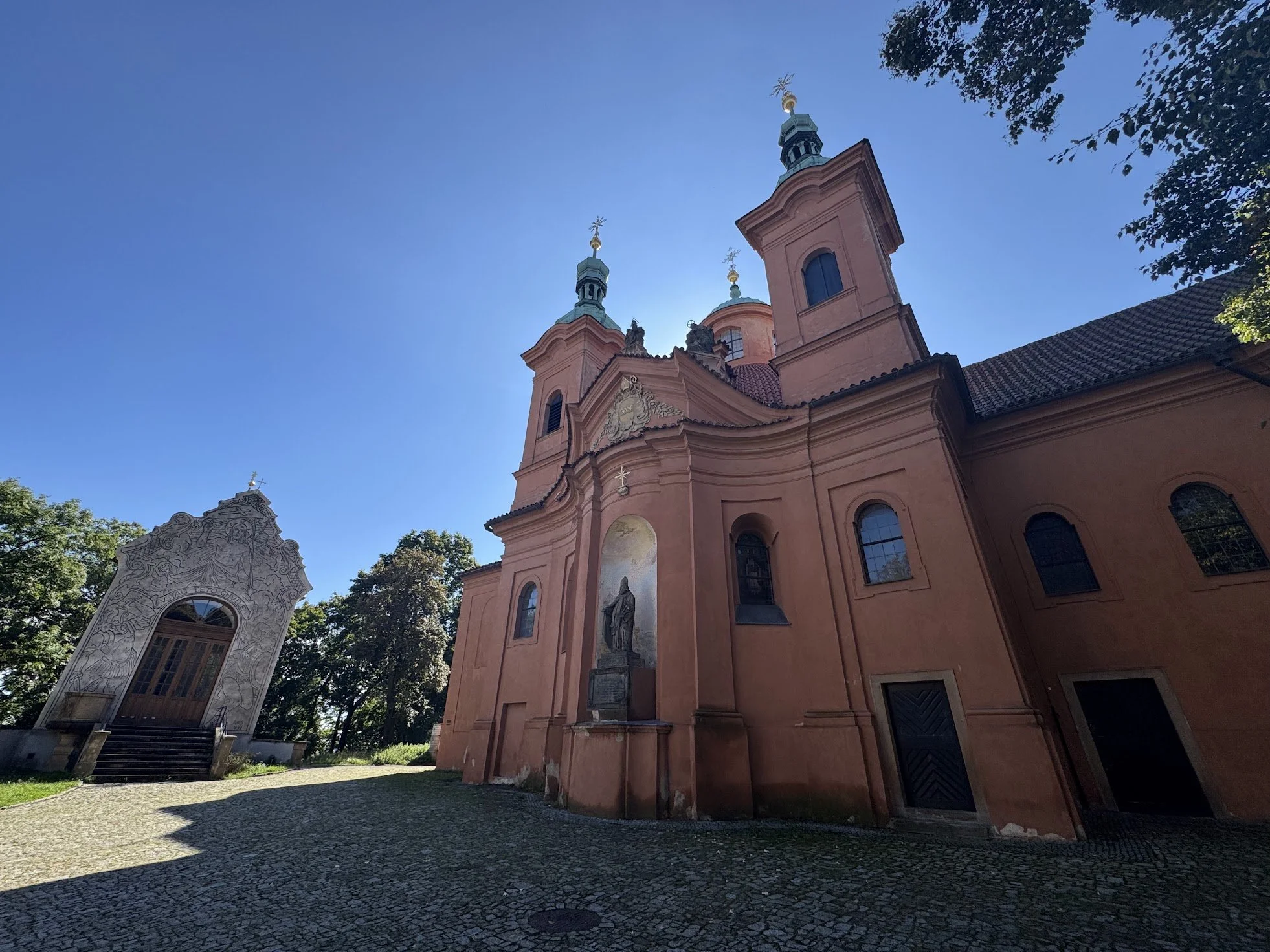 A pink church with two bell towers topped with green spires and gold crosses, surrounded by trees, with a clear blue sky overhead.