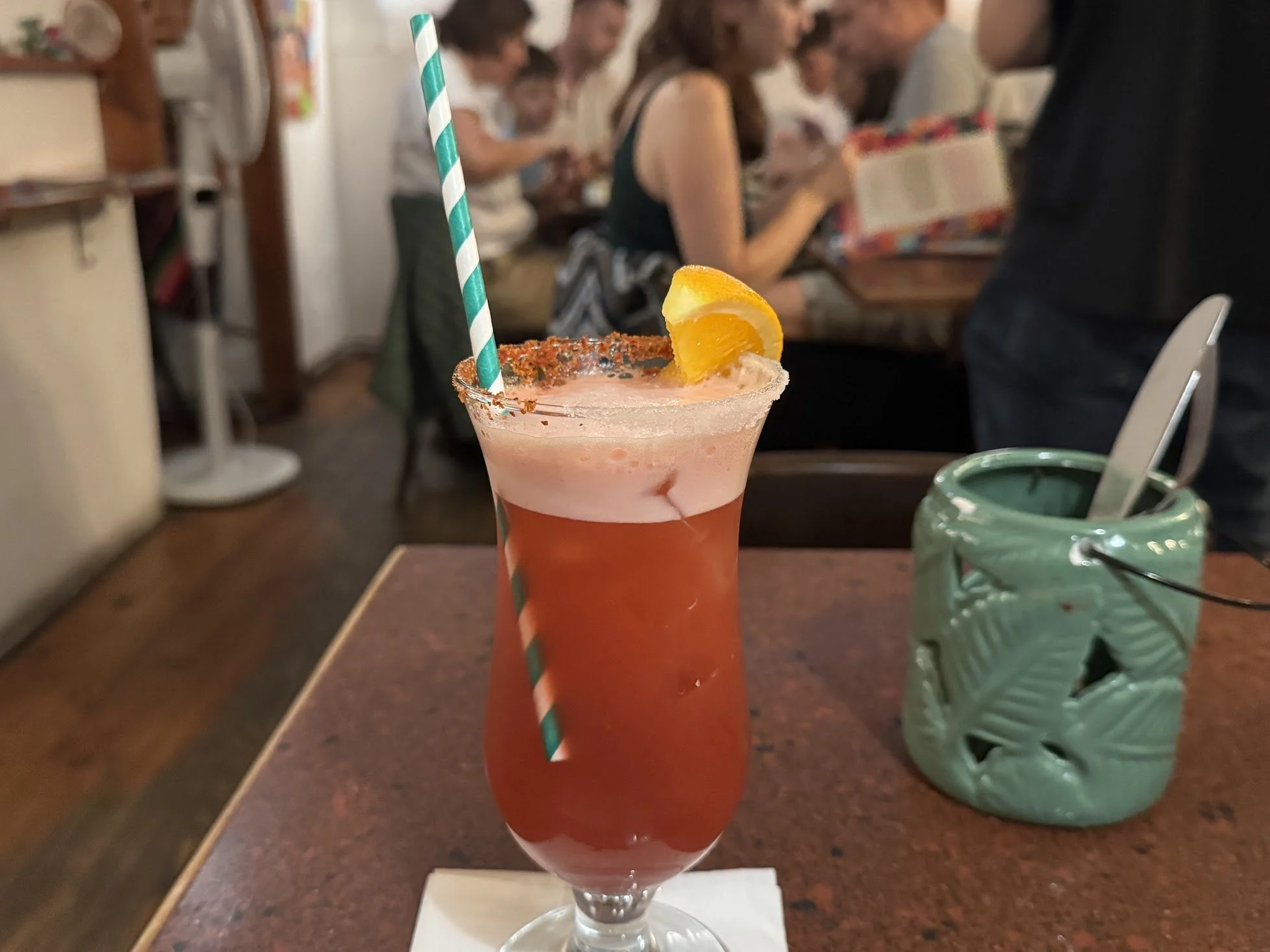 A pink cocktail drink garnished with a lemon wedge and a blue and white striped paper straw, on a restaurant table with people in the background.