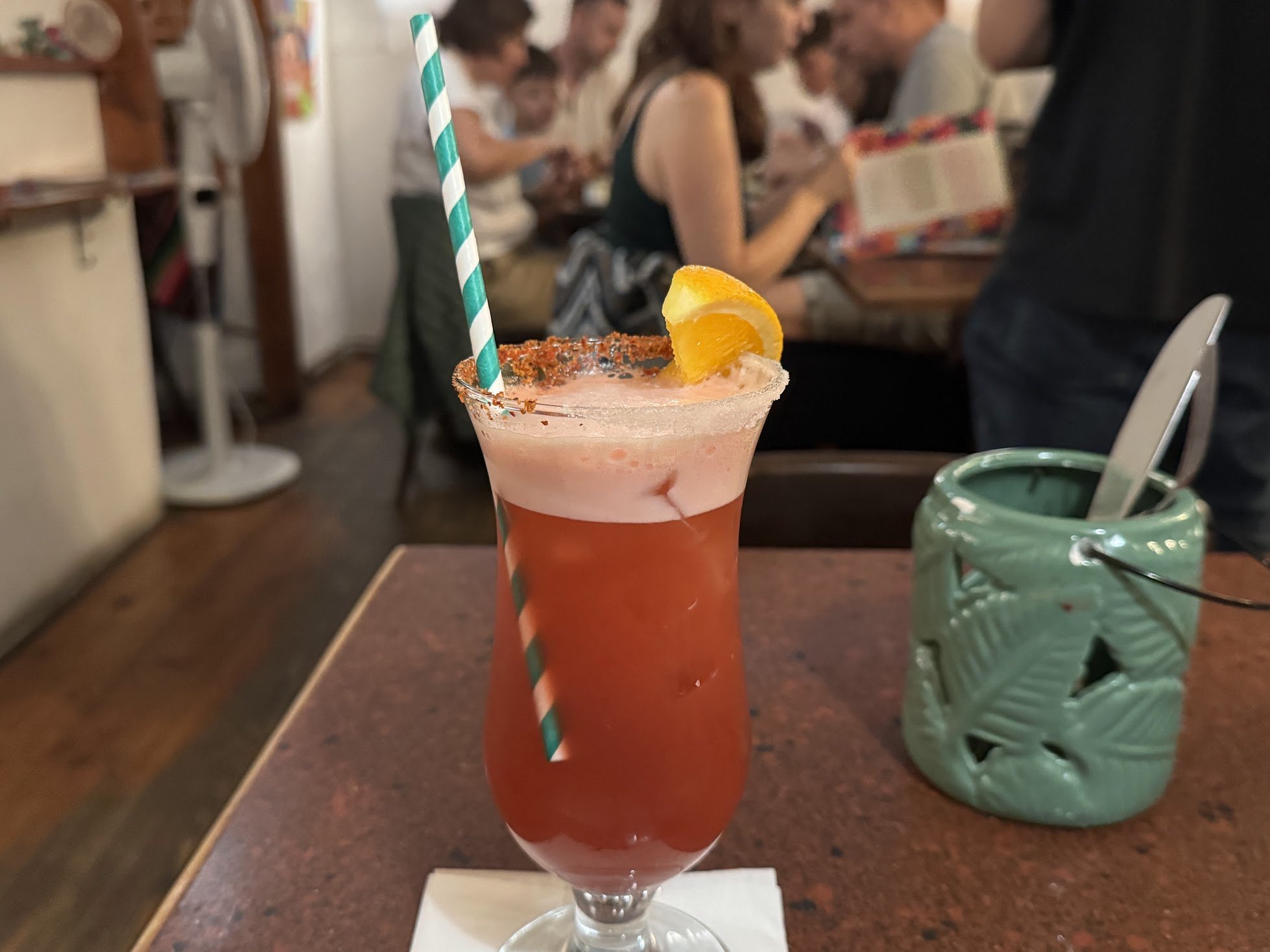 A pink cocktail drink with an orange wedge garnish and a teal-and-white striped straw on a table inside a lively restaurant, with people reading menus in the background.