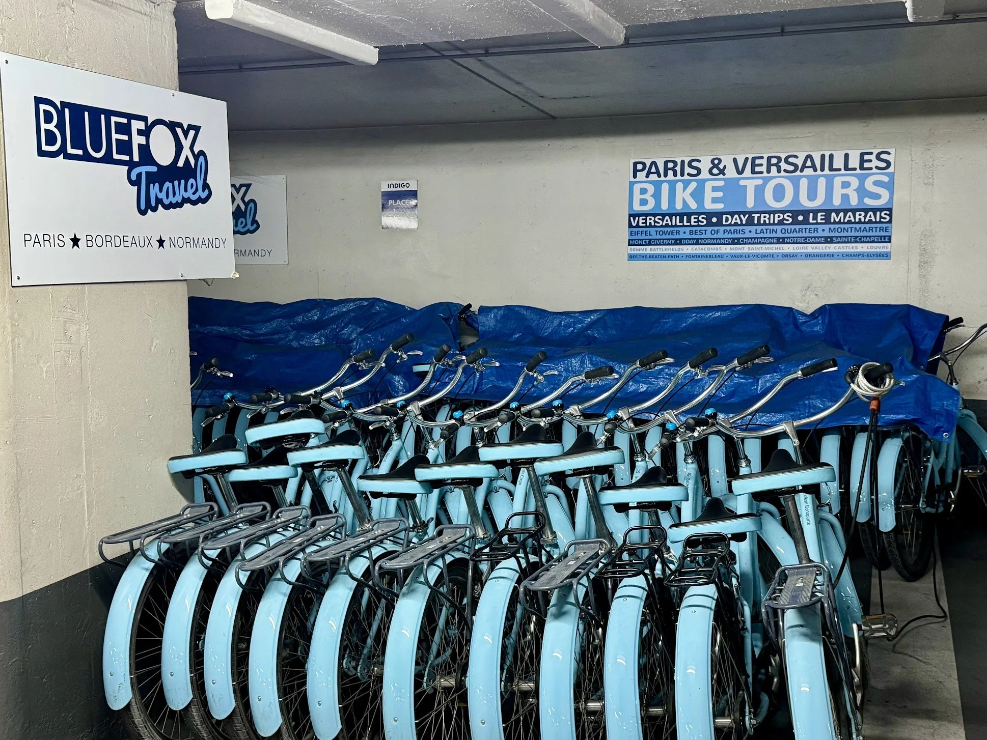 A row of light blue bicycles parked and locked under a covered area with signs indicating bike tours in Paris and Versailles, France.