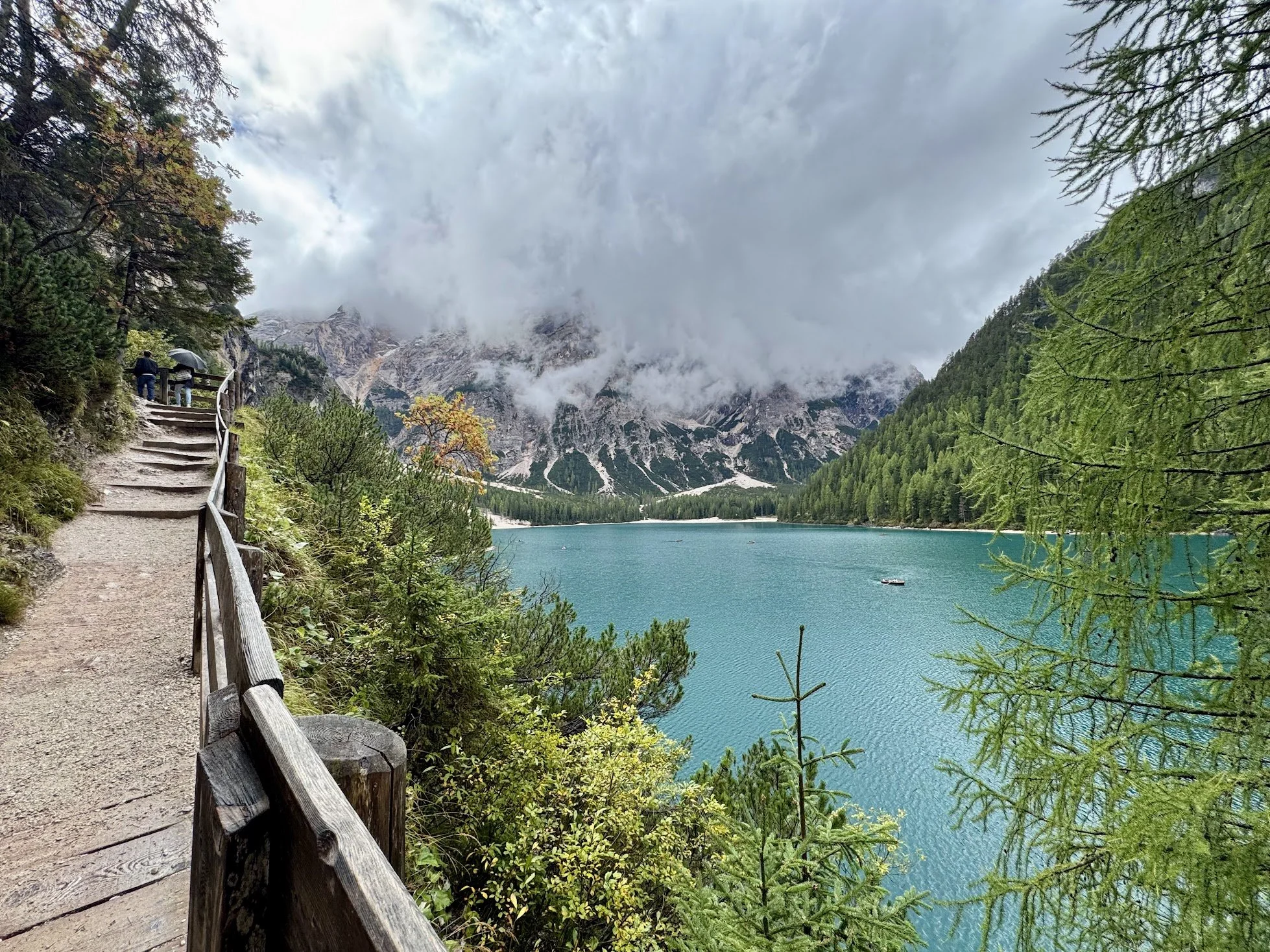 A lakeside hiking trail with wooden railing, surrounded by green trees, leading towards a mountain range with clouds overhead and a boat on the lake.